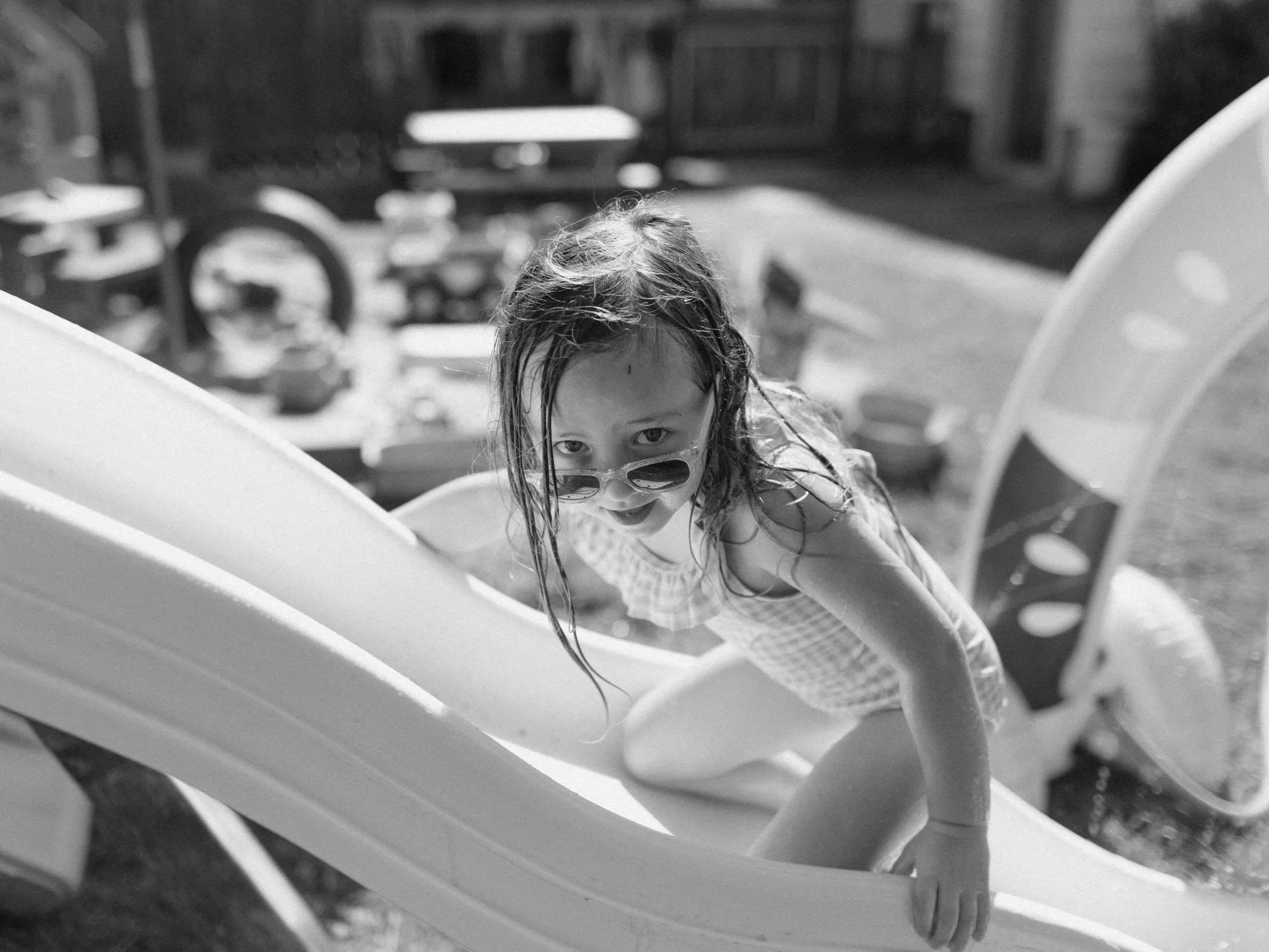 A young girl with wet hair wearing sunglasses and a checkered swimsuit is climbing on a slide in a backyard. The background shows a sandbox, toy wagon, and outdoor furniture.