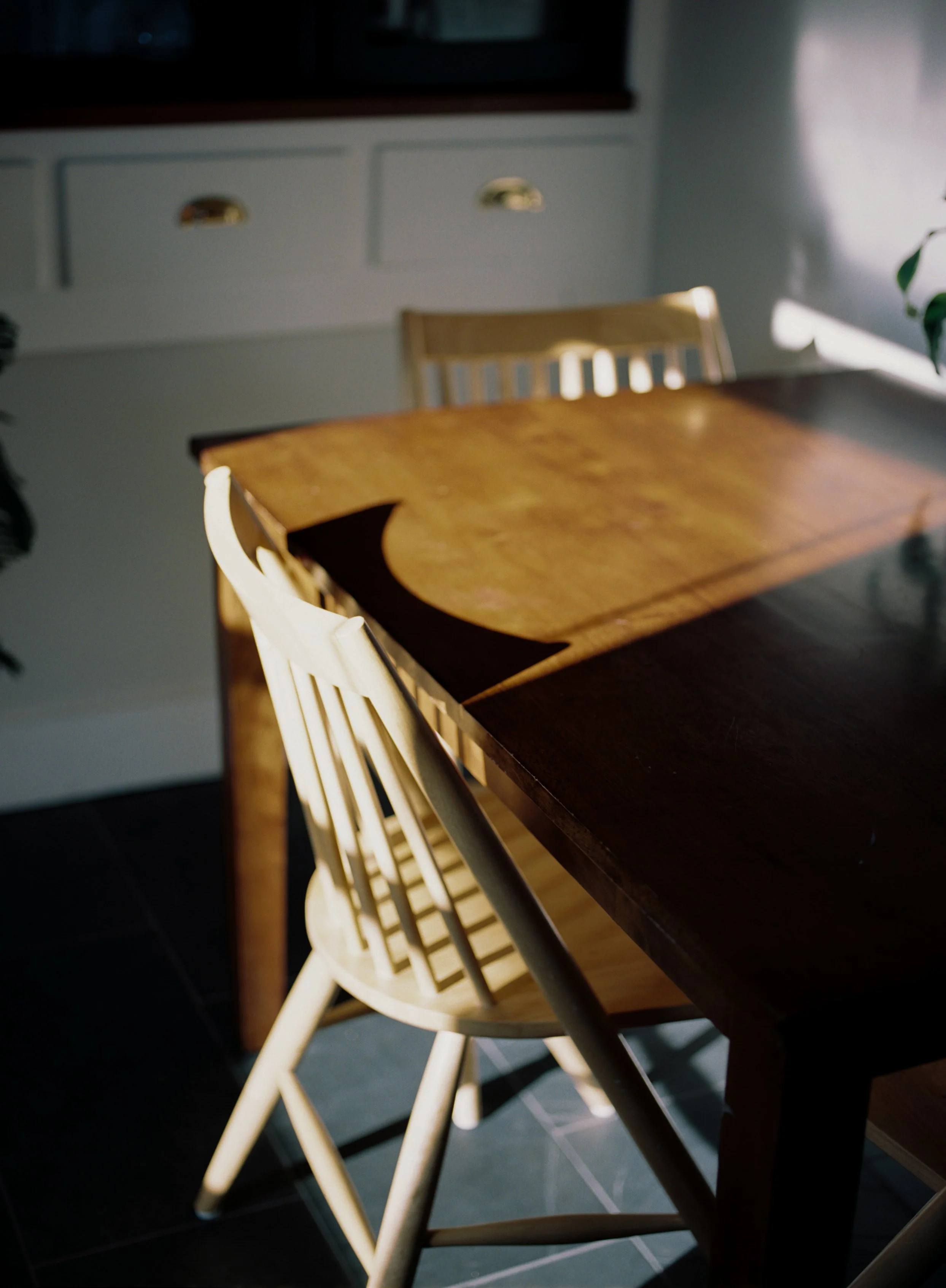 A wooden dining table with sunlight casting shadows on it, surrounded by light-colored wooden chairs, in a room with white walls and cabinets.