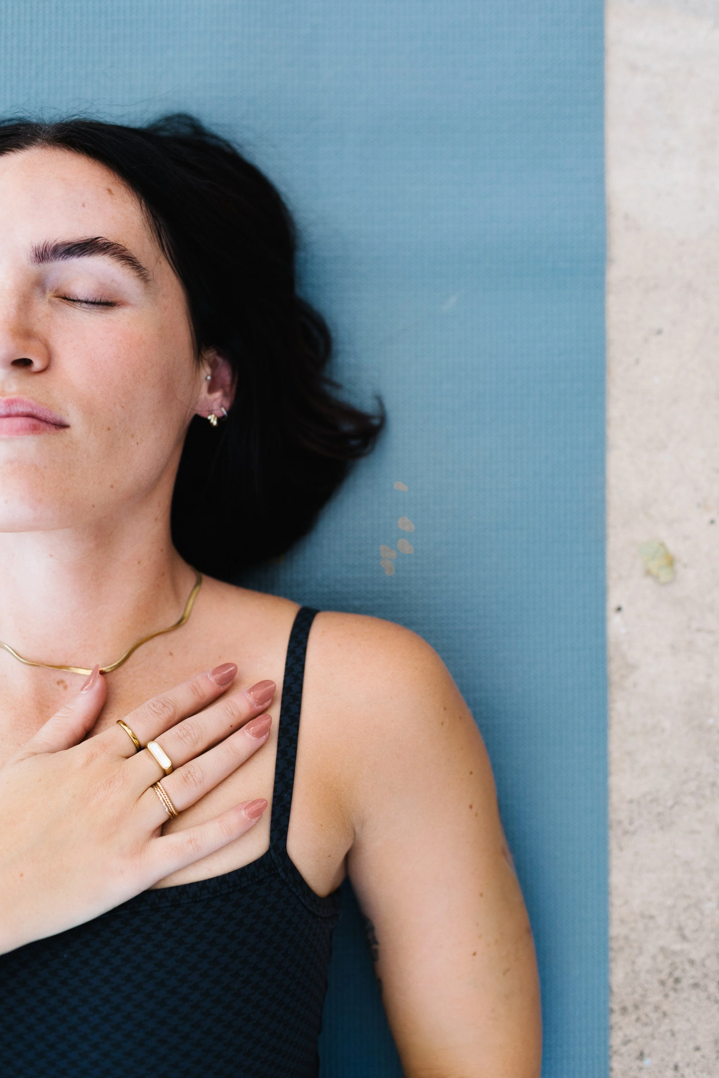 A woman with dark hair and fair skin, lying on a blue yoga mat with her eyes closed, dressed in a black patterned tank top, wearing gold jewelry, and resting her hand on her chest.