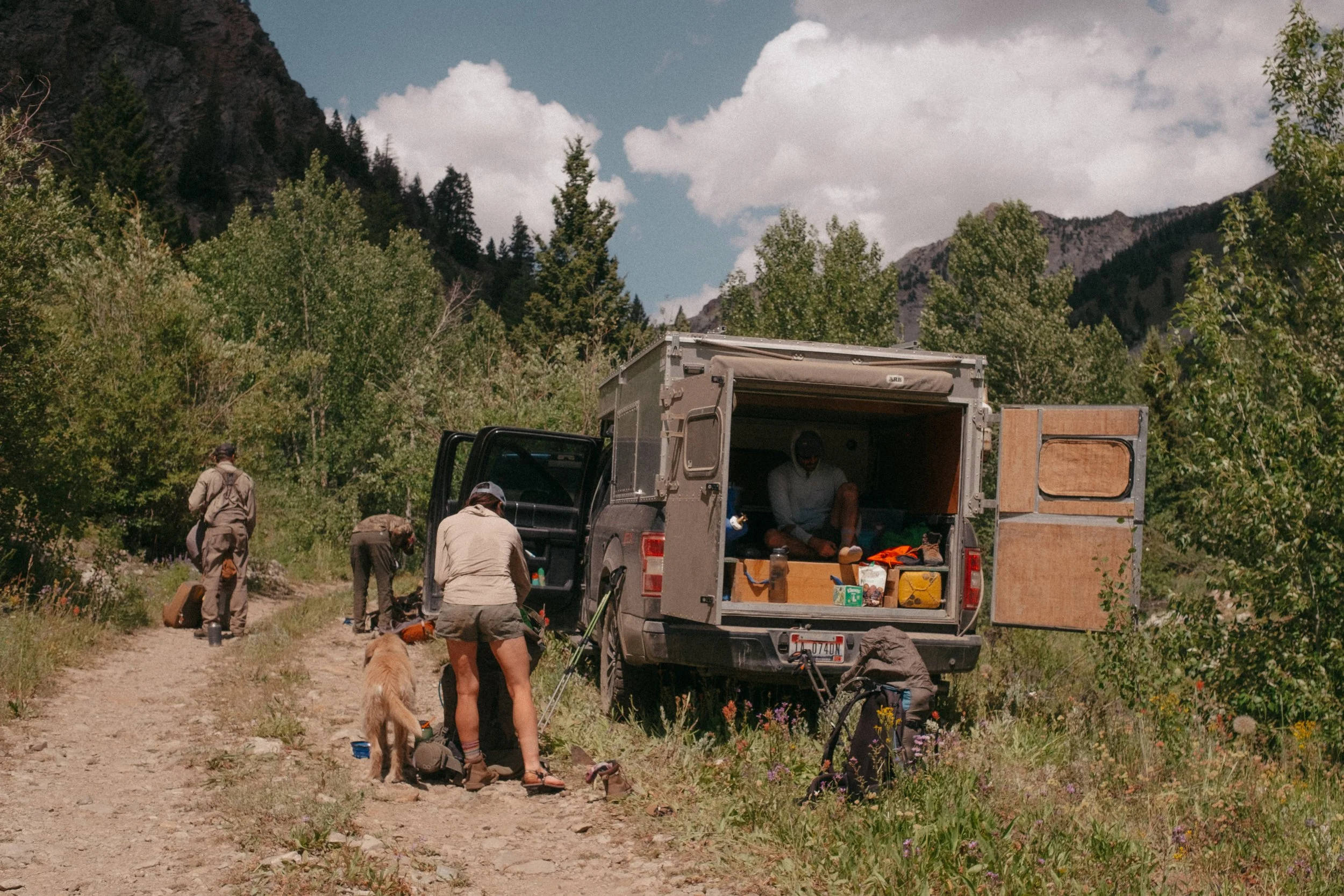 People camping in a mountain forest, with a van and truck parked on a dirt trail, surrounded by green trees and mountains under a partly cloudy sky.