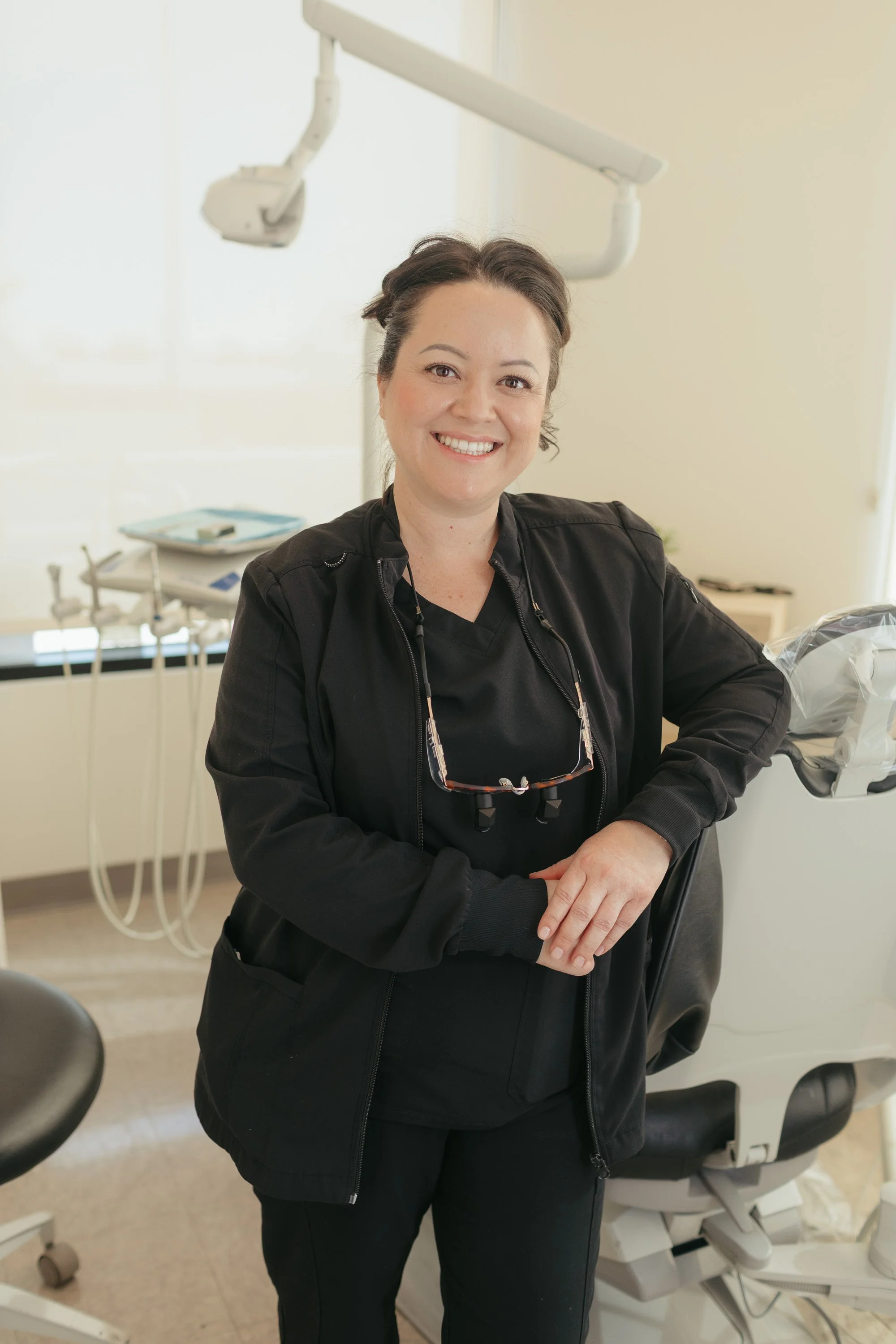 Smiling woman in black scrubs standing in dental office with dental chair and equipment.