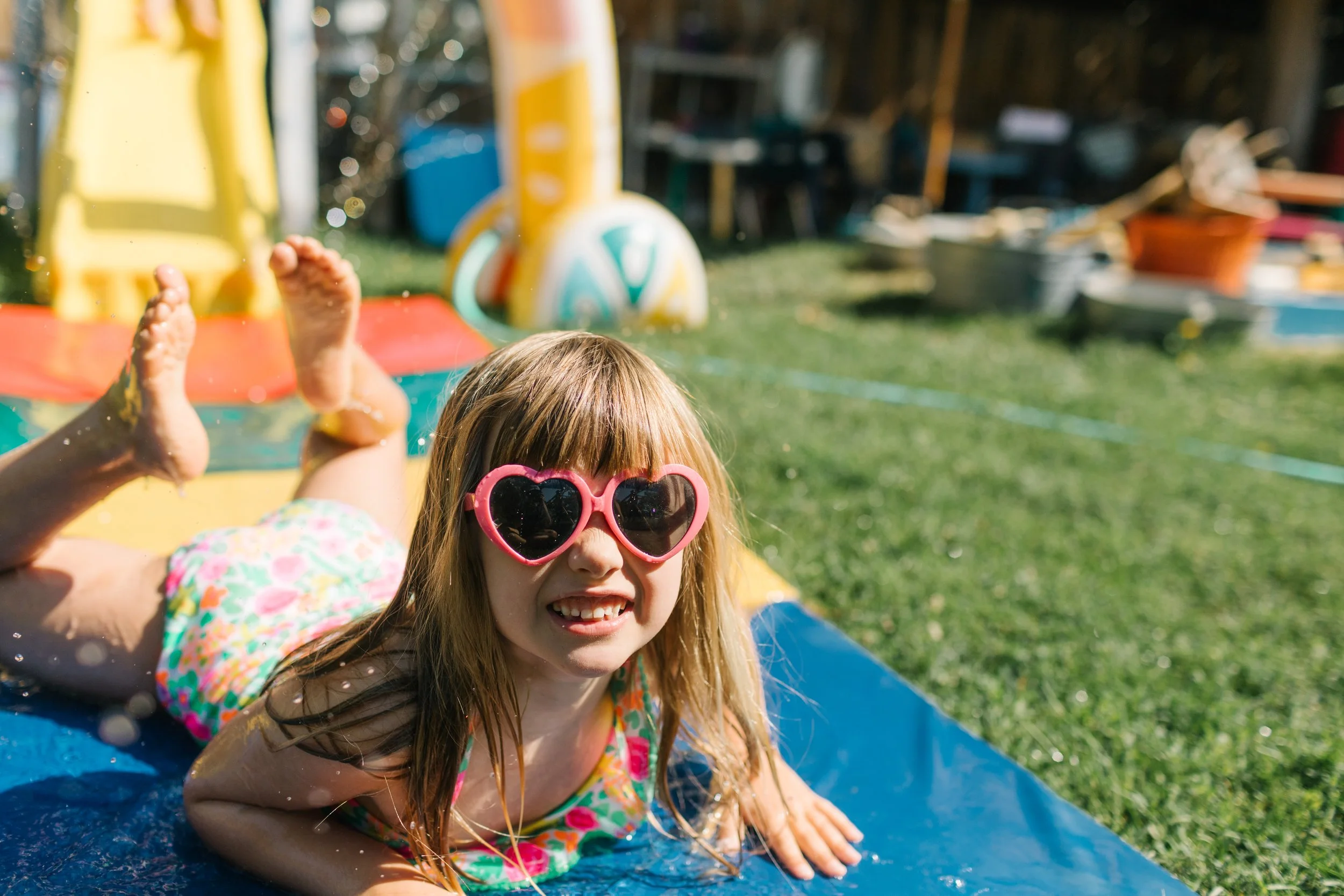 A girl with long hair and heart-shaped sunglasses smiling, lying on a slip and slide in a backyard, with water splashing around her, in sunny weather.