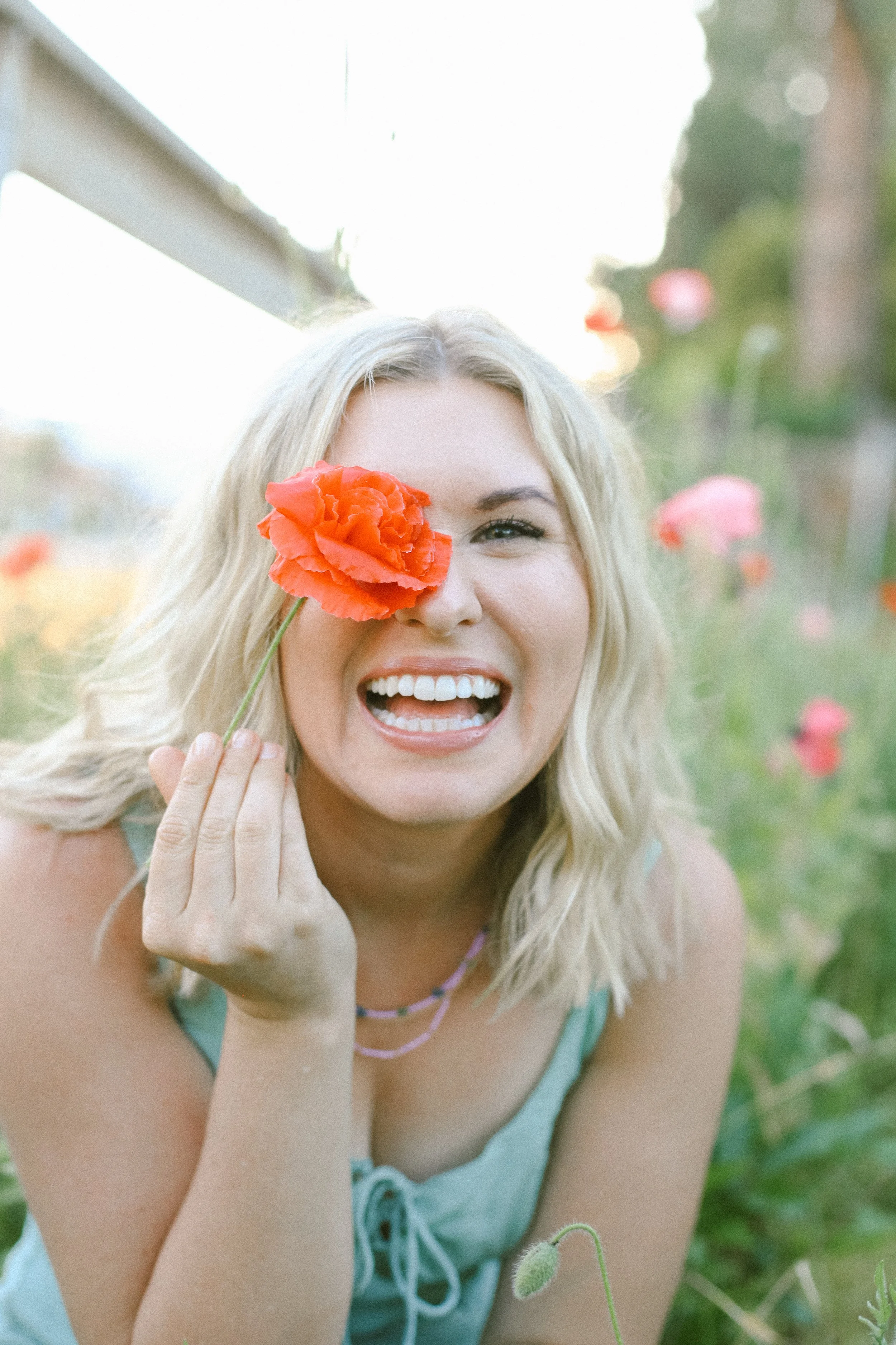 A young woman with blonde hair, smiling and holding an orange flower over one eye, surrounded by a garden of pink and orange flowers.