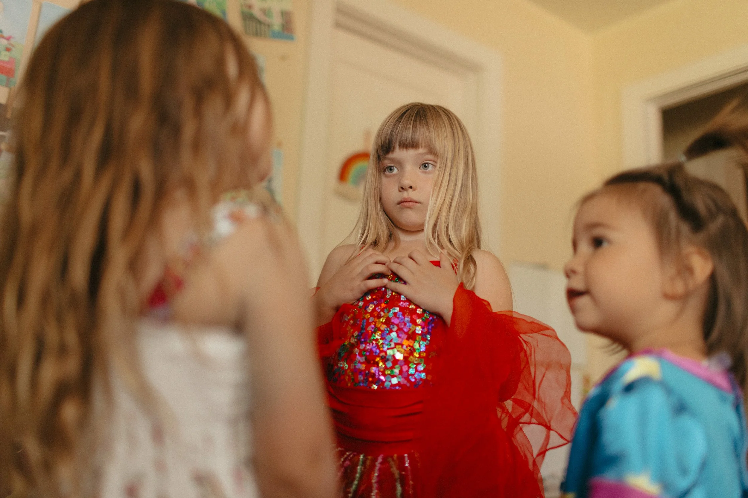 Three young girls having a conversation indoors; one girl in the background has blonde hair and is wearing a red outfit with sequins, looking surprised or concerned, while the other two girls in the foreground have brown hair and are wearing colorful