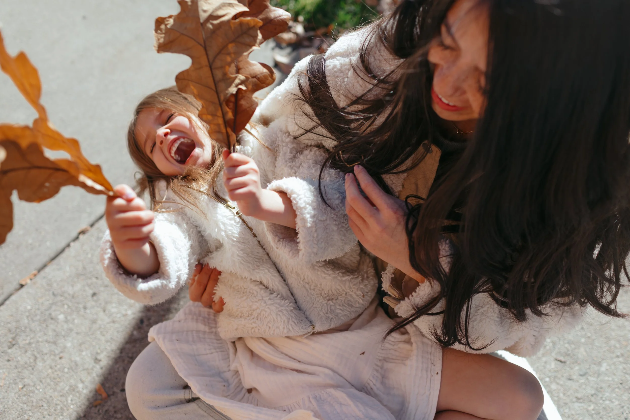 A woman and a young girl playing outside in autumn in Boise, Idaho.. The girl is laughing and holding large brown leaves, while the woman smiles at her.