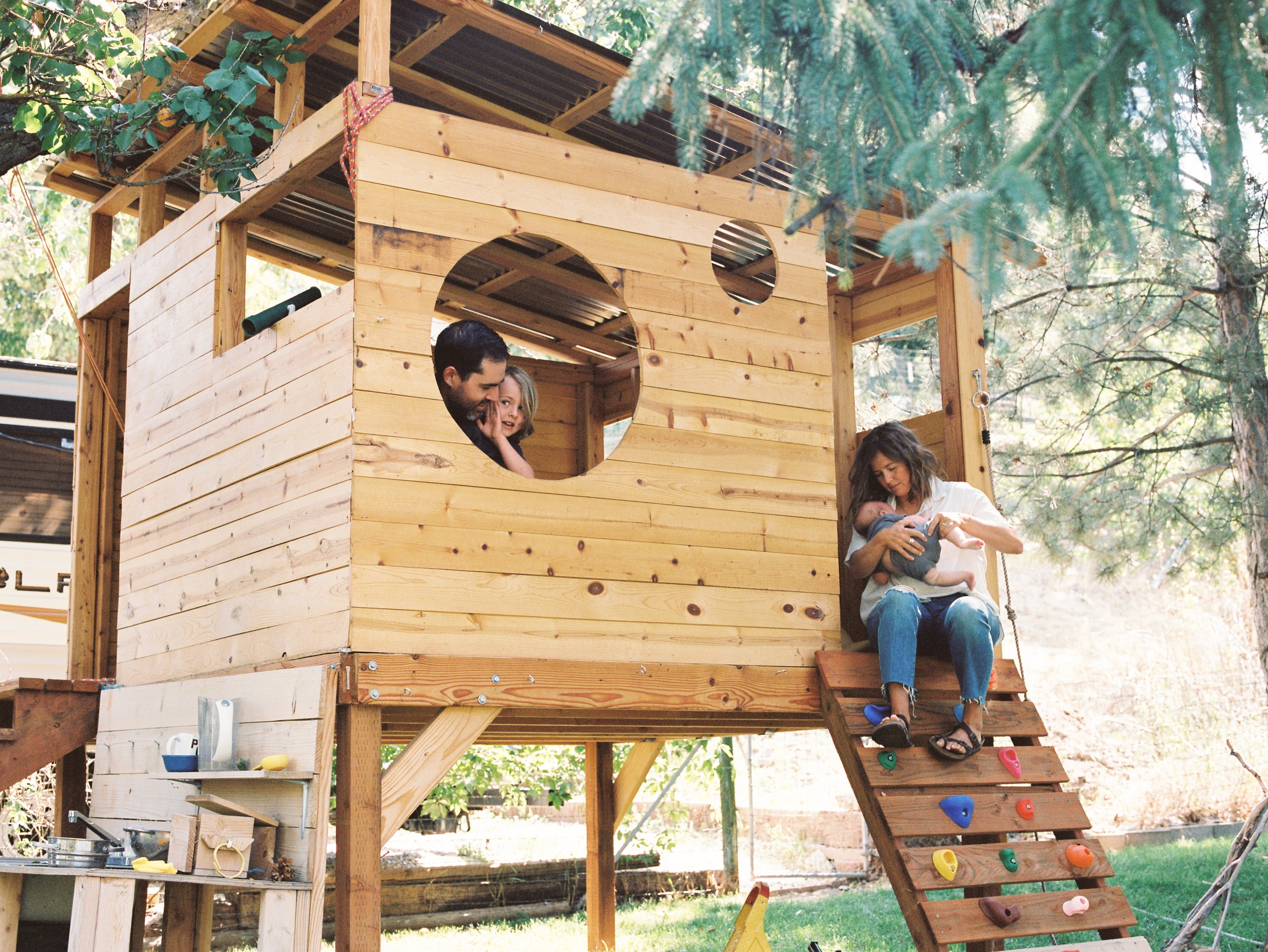 Family enjoying time in their backyard treehouse in Boise, Idaho, with children peering out the window and a woman sitting on the steps holding a baby.