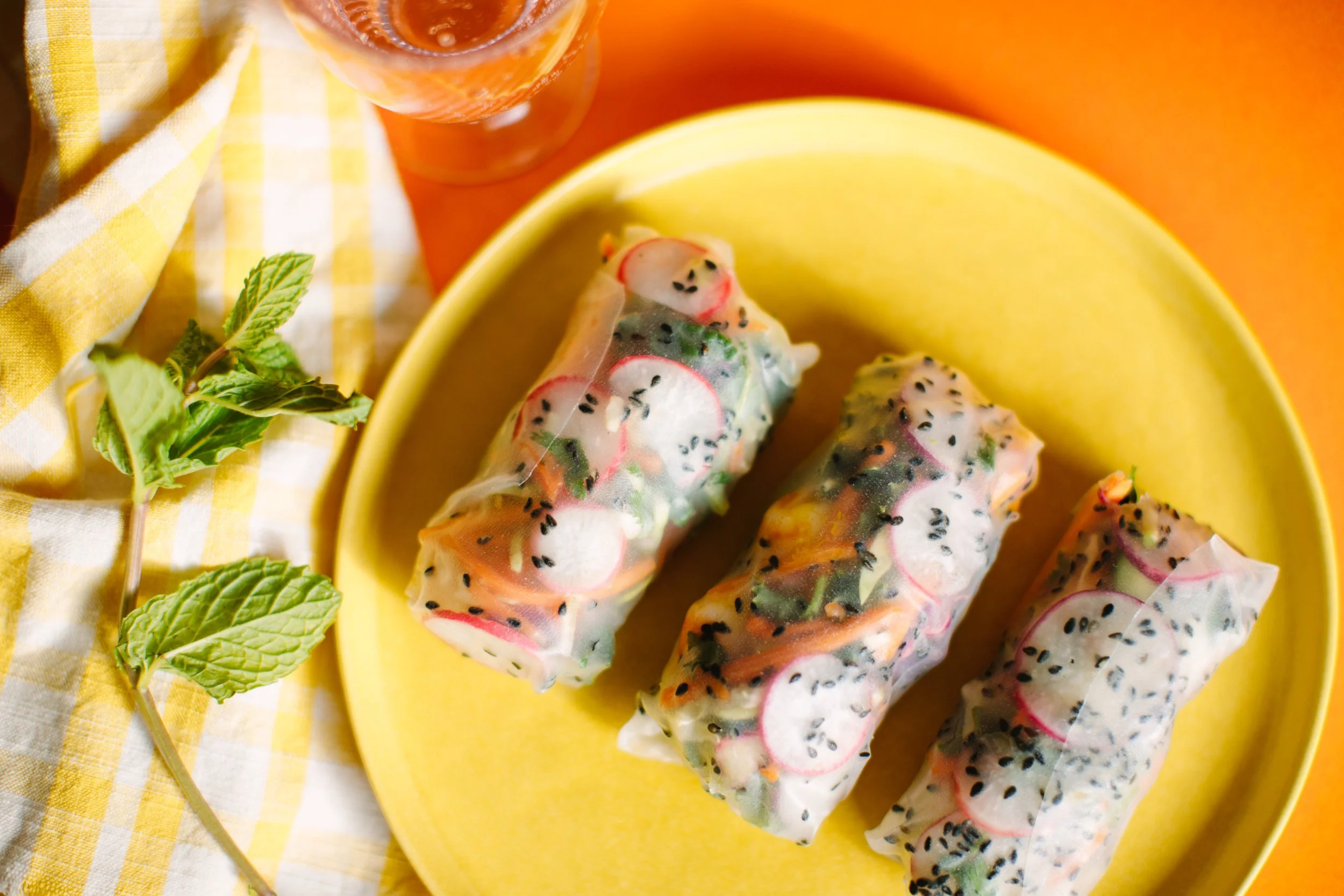 Three fresh spring rolls filled with sliced radishes, carrots, and greens on a yellow plate. A sprig of mint and a glass of iced beverage are beside the plate, with a yellow and white checkered cloth nearby.