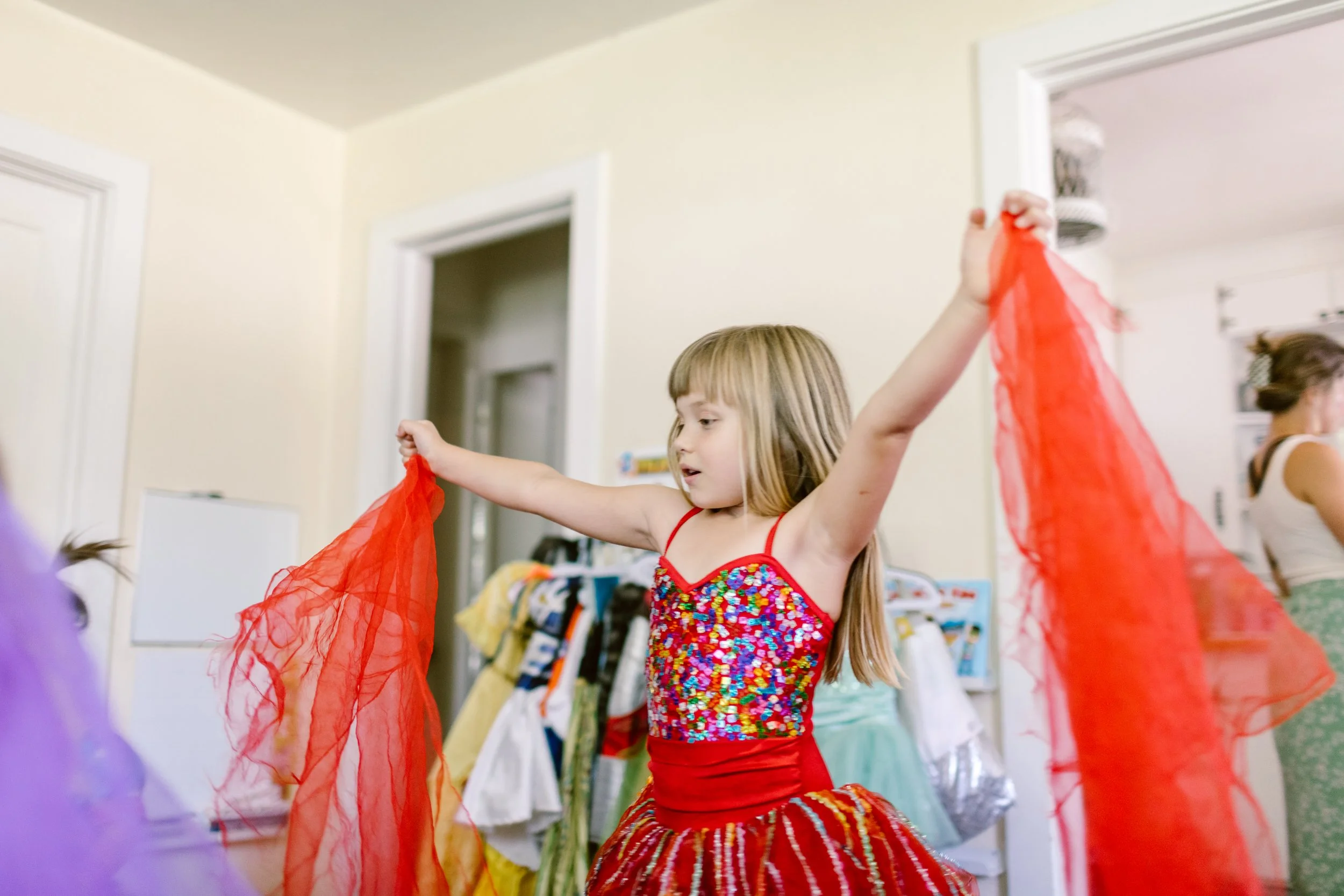 A young girl with long blonde hair wearing a colorful, sparkly dress and a red tutu dancing with red and purple fabric scarves in a room filled with clothes.