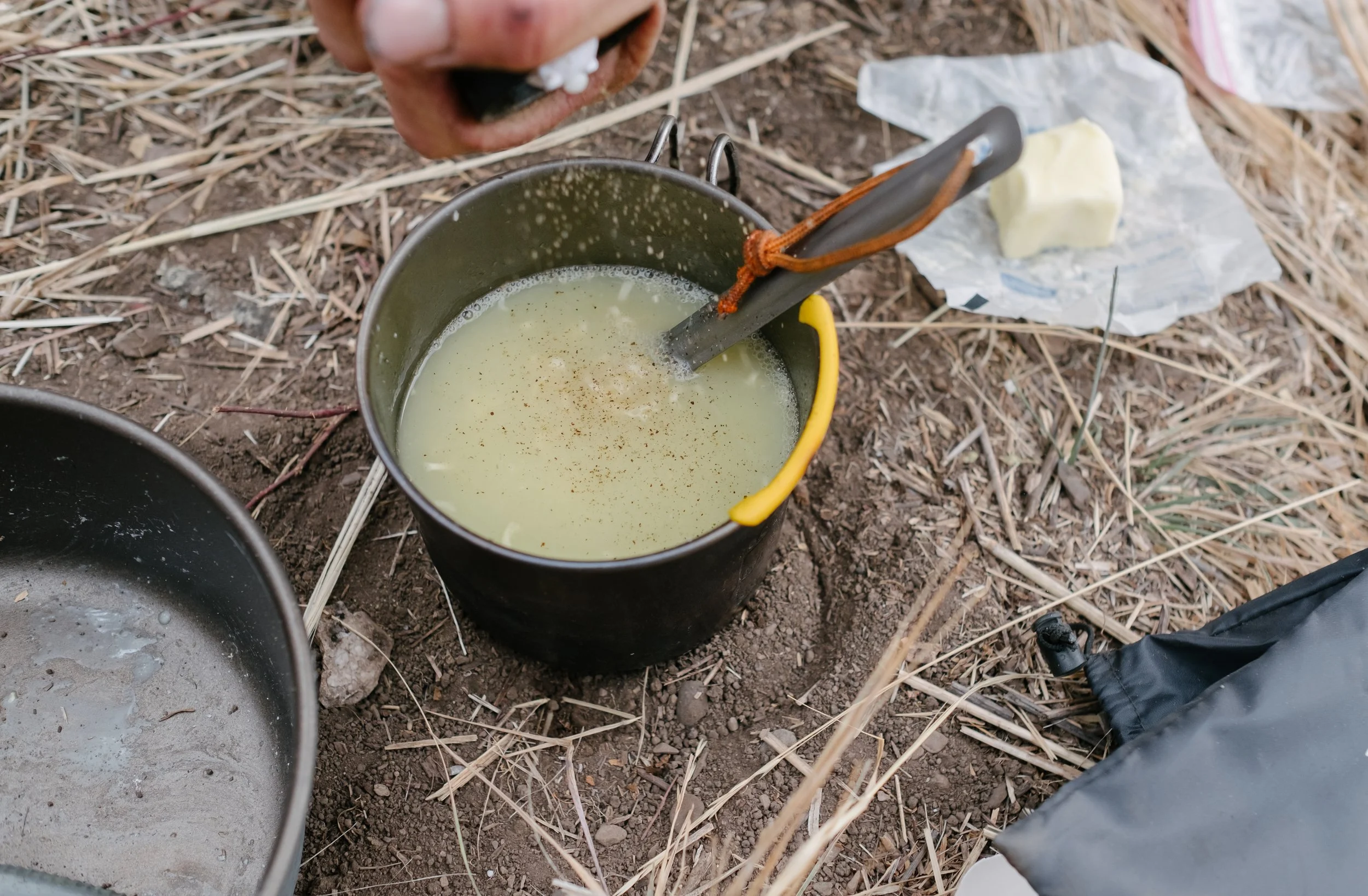 A camping scene showing a pot of melted butter on the ground, with a spoon inside, surrounded by dry grass and a block of butter on paper nearby.