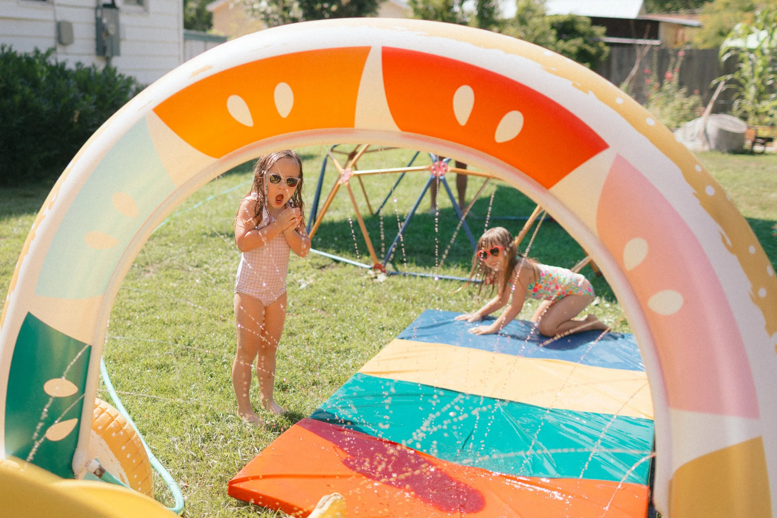 Two young girls playing with a splash pad indoors during summer; one girl is standing with water on her face, wearing sunglasses and a pink swimsuit, the other is on her hands and knees on a colorful mat, also wearing sunglasses and a swimsuit; the s