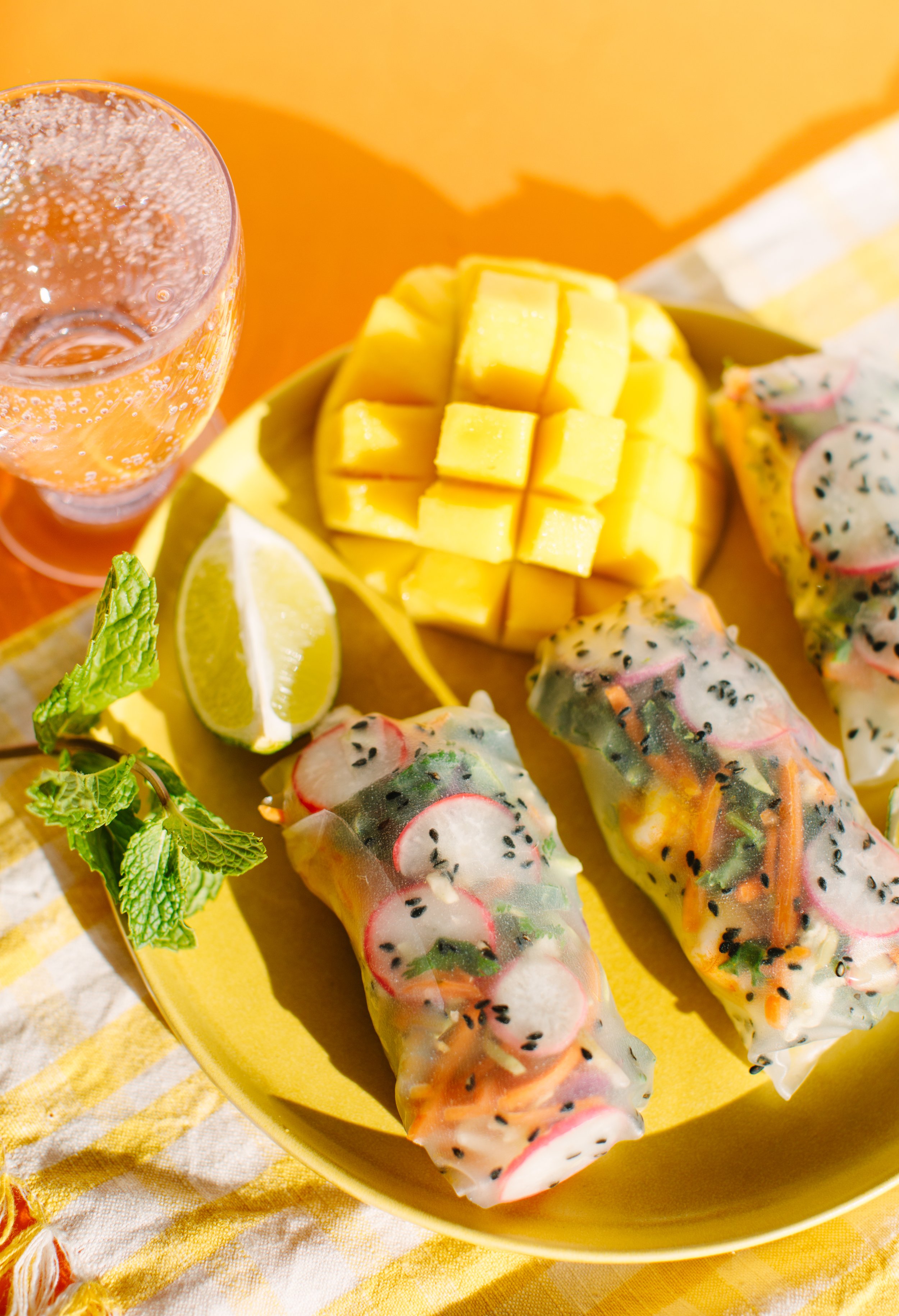 Fresh spring rolls filled with vegetables and herbs on a yellow plate, with sliced radishes, mango chunks, and lime wedges, accompanied by a glass of sparkling beverage garnished with mint.