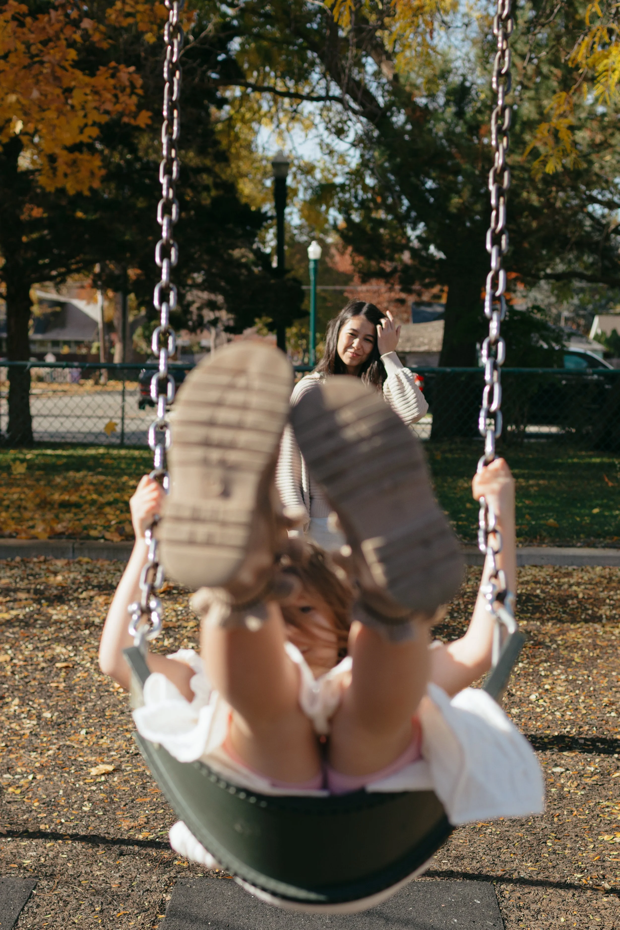 A woman standing behind a child on a swing in a park during fall, with trees and autumn leaves in the background.