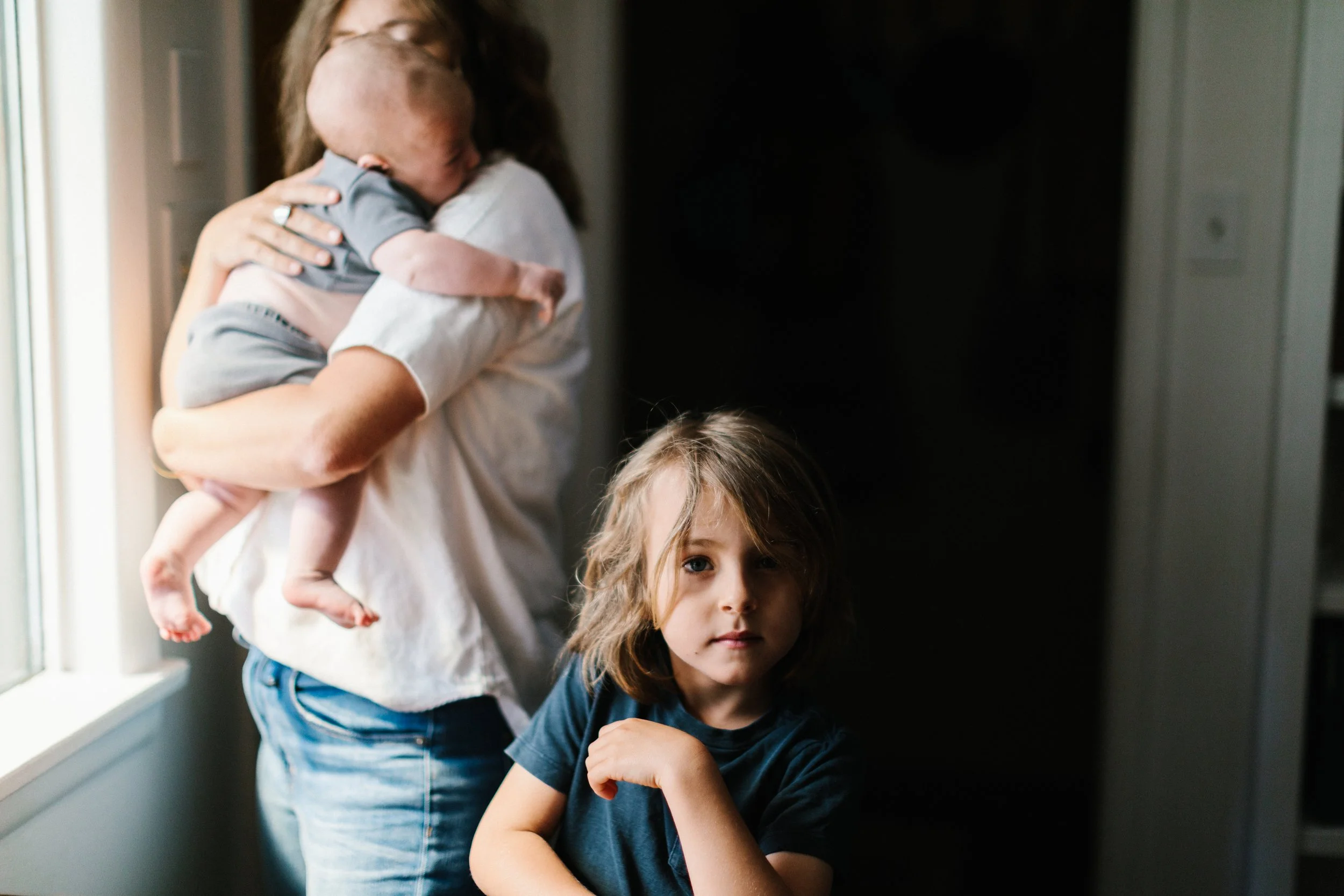 A woman holds a baby while a young girl with shoulder-length hair looks at the camera inside a room near a window, with a dark background behind them.