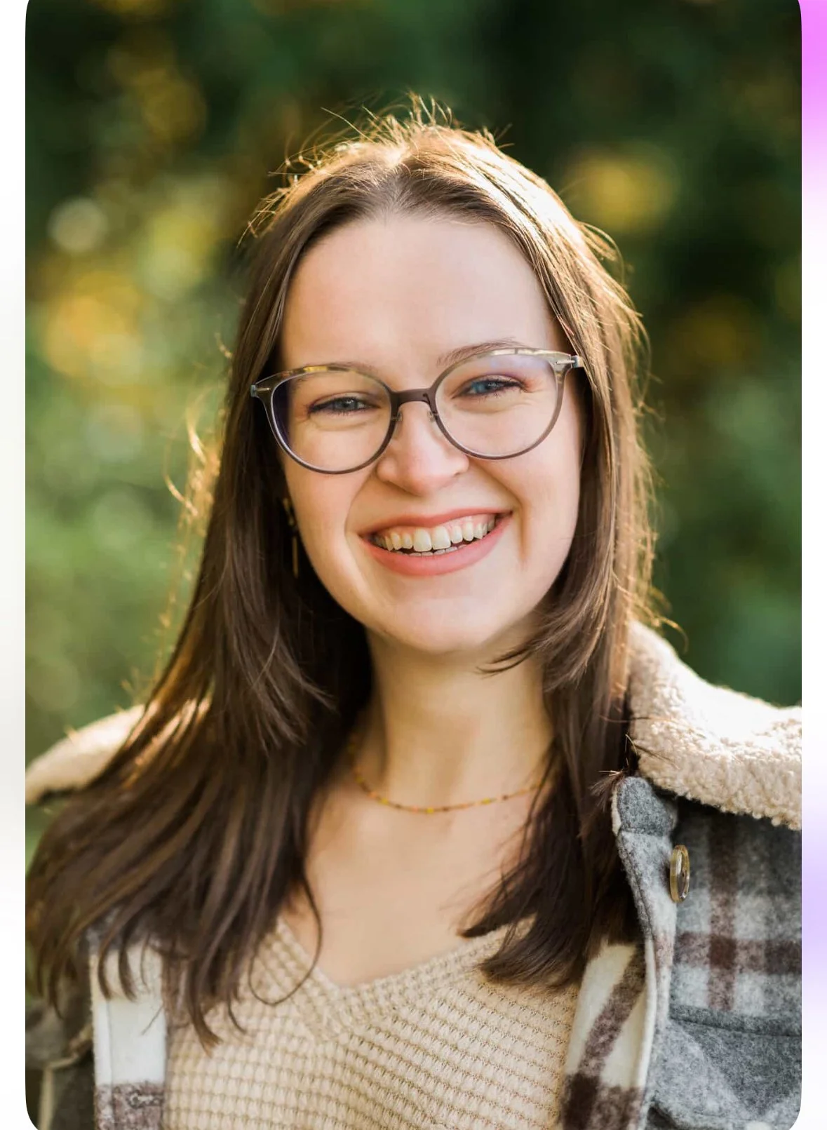 A young woman with long brown hair, glasses, and a warm smile, standing outdoors with greenery in the background.