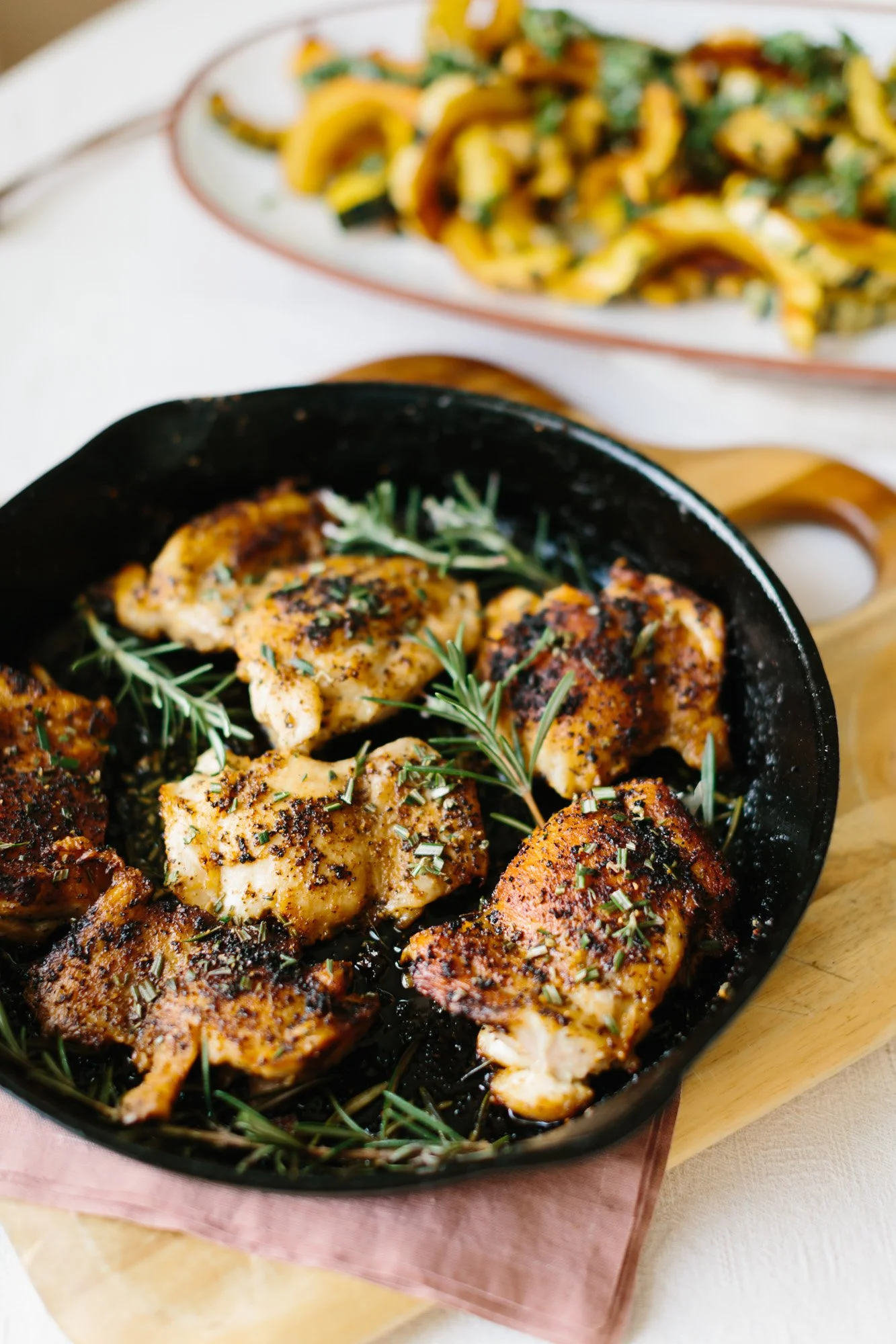 Cooked seasoned chicken pieces garnished with herbs in a black skillet on a wooden board, with a plate of cooked vegetables in the background.