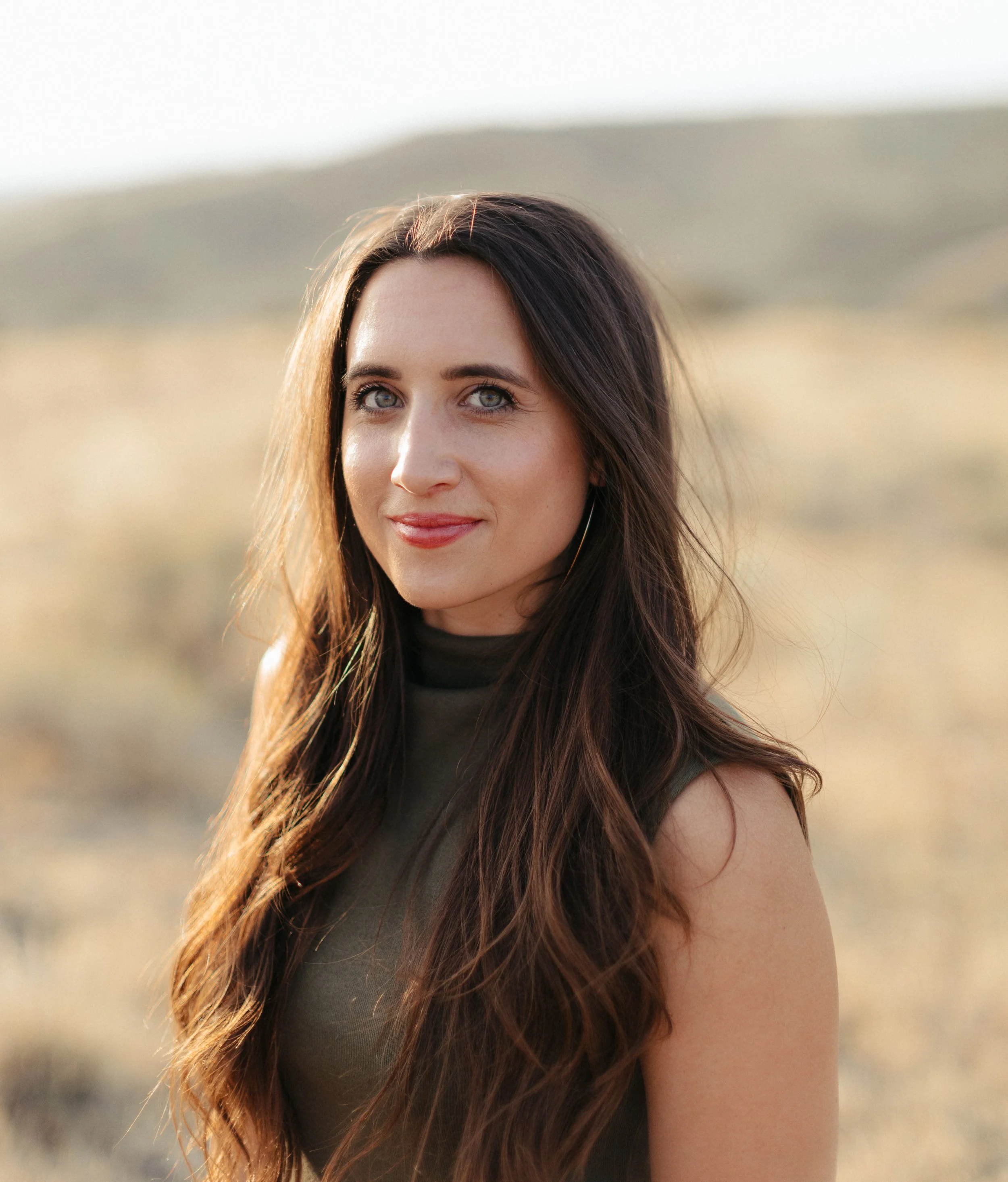 A woman with long brown hair and blue eyes stands outdoors in a desert landscape during sunset.