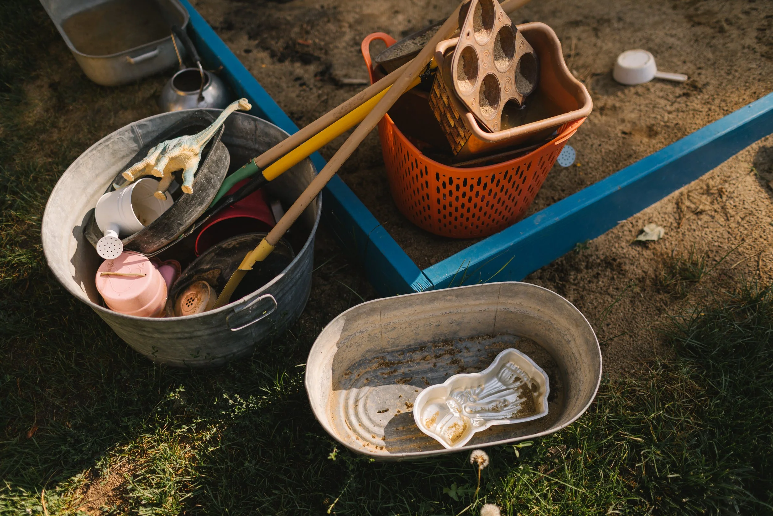 Child's sandbox with various plastic toys, tools, and containers, some dirty and wet, outside in the grass and dirt.