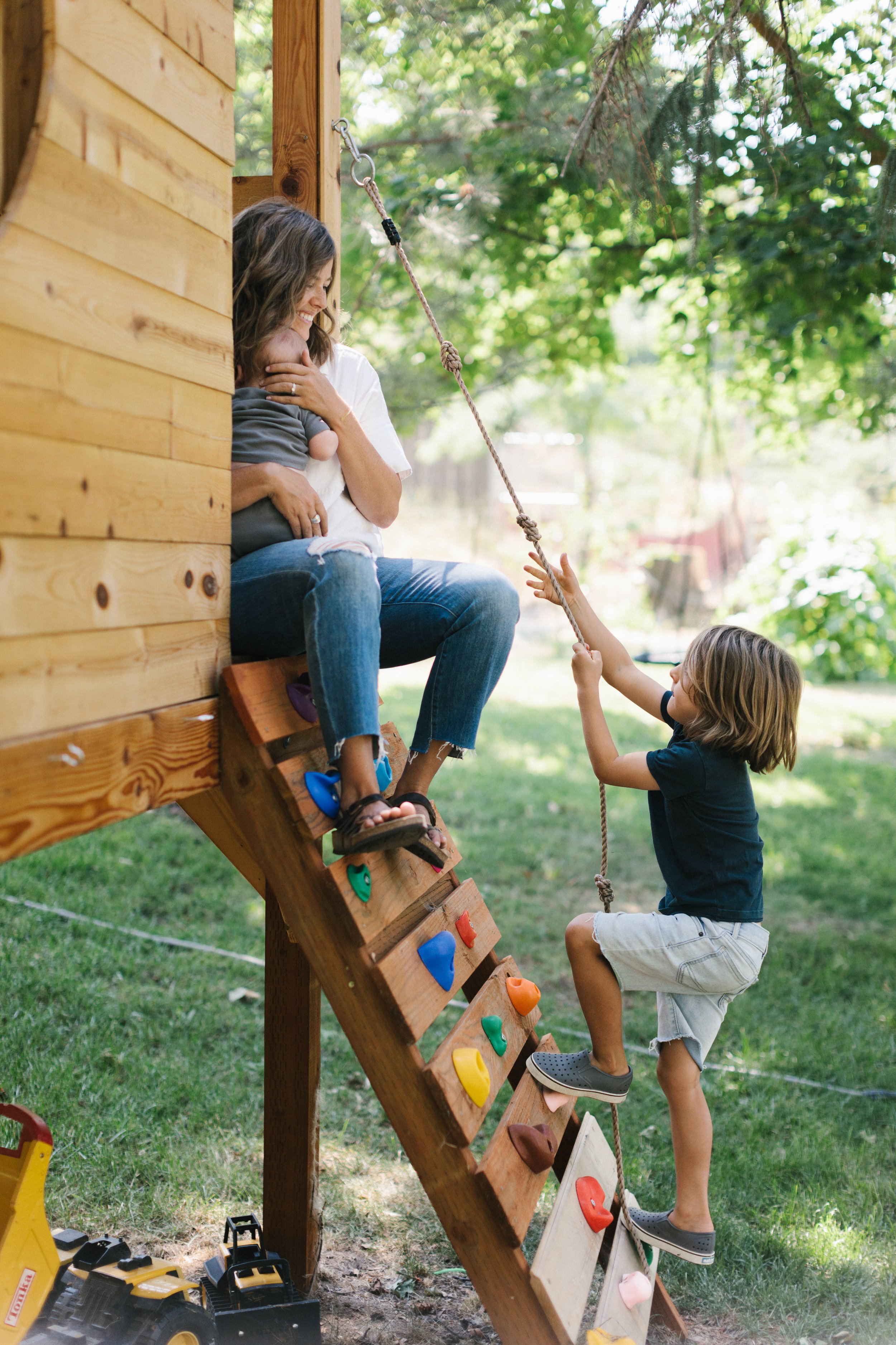 A woman sitting on a wooden playhouse staircase outside, holding a young child, while a boy climbs the stairs reaching towards her, in a backyard with grass and trees. Image taken by Boise Lifestyle family photographer.