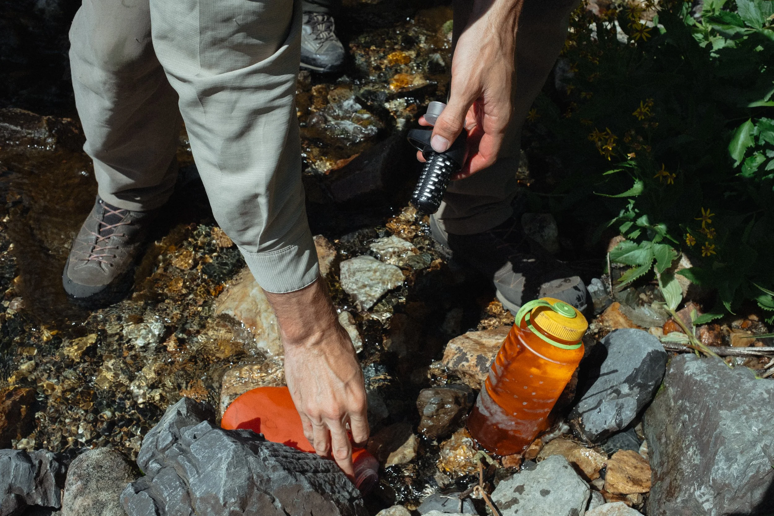 Person filling a plastic water bottle from a small mountain stream using a filter pump, with other bottles nearby, surrounded by rocks and greenery.