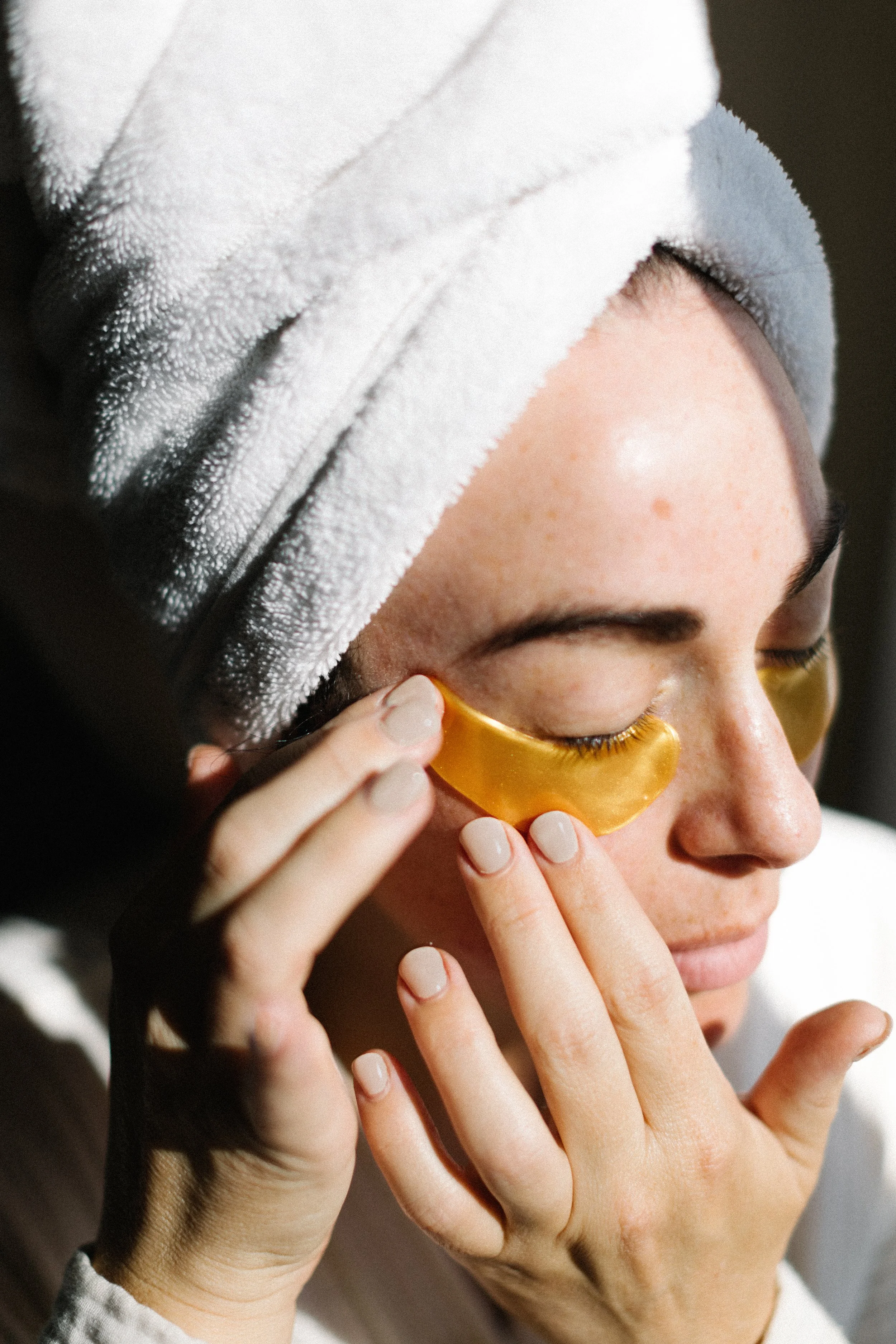 Close-up of a woman with a white towel on her head, gently pressing a gold under-eye patch onto her closed eye with her index finger.