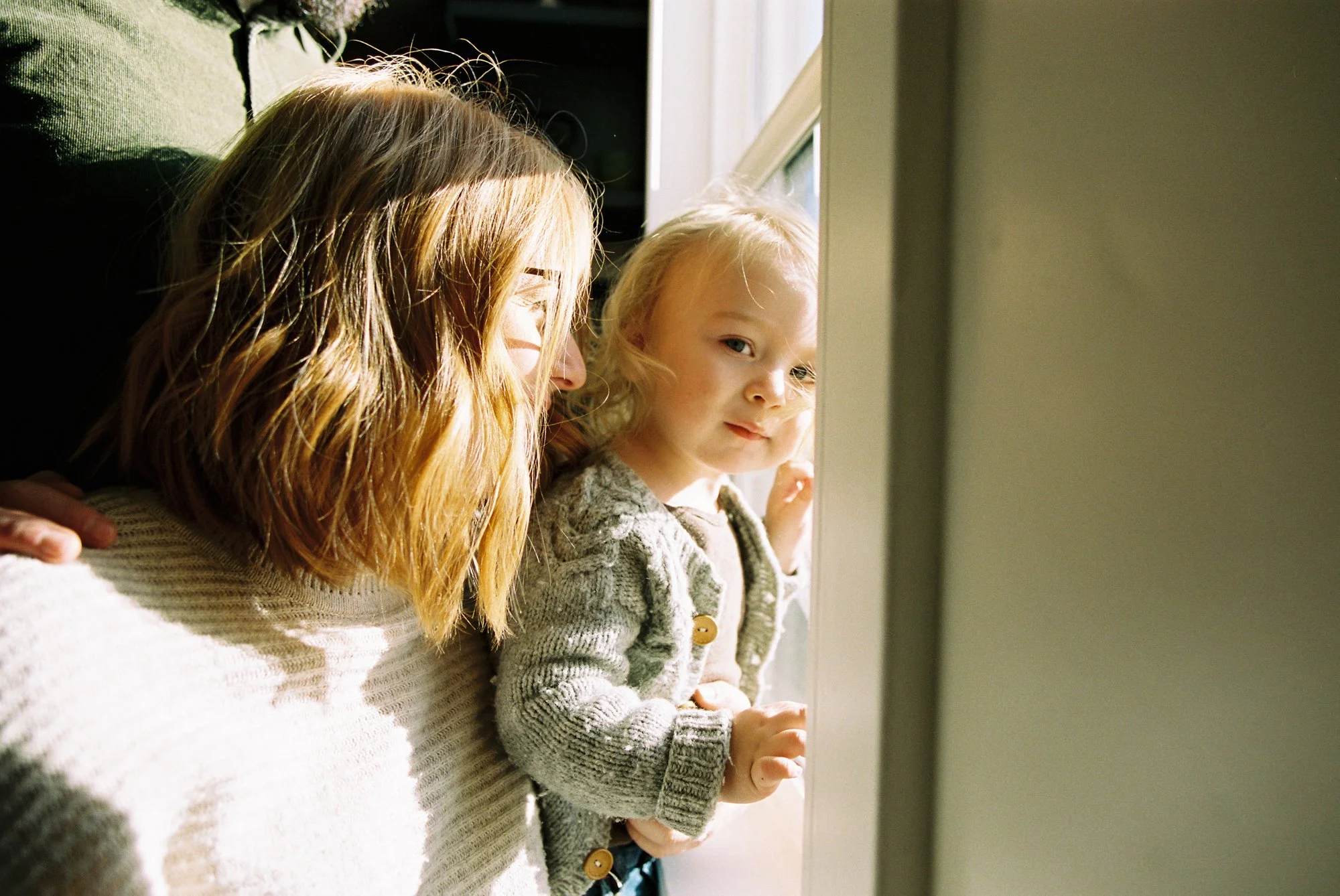 A woman with red hair holding a young girl with blonde hair looking out a window with sunlight streaming in.
