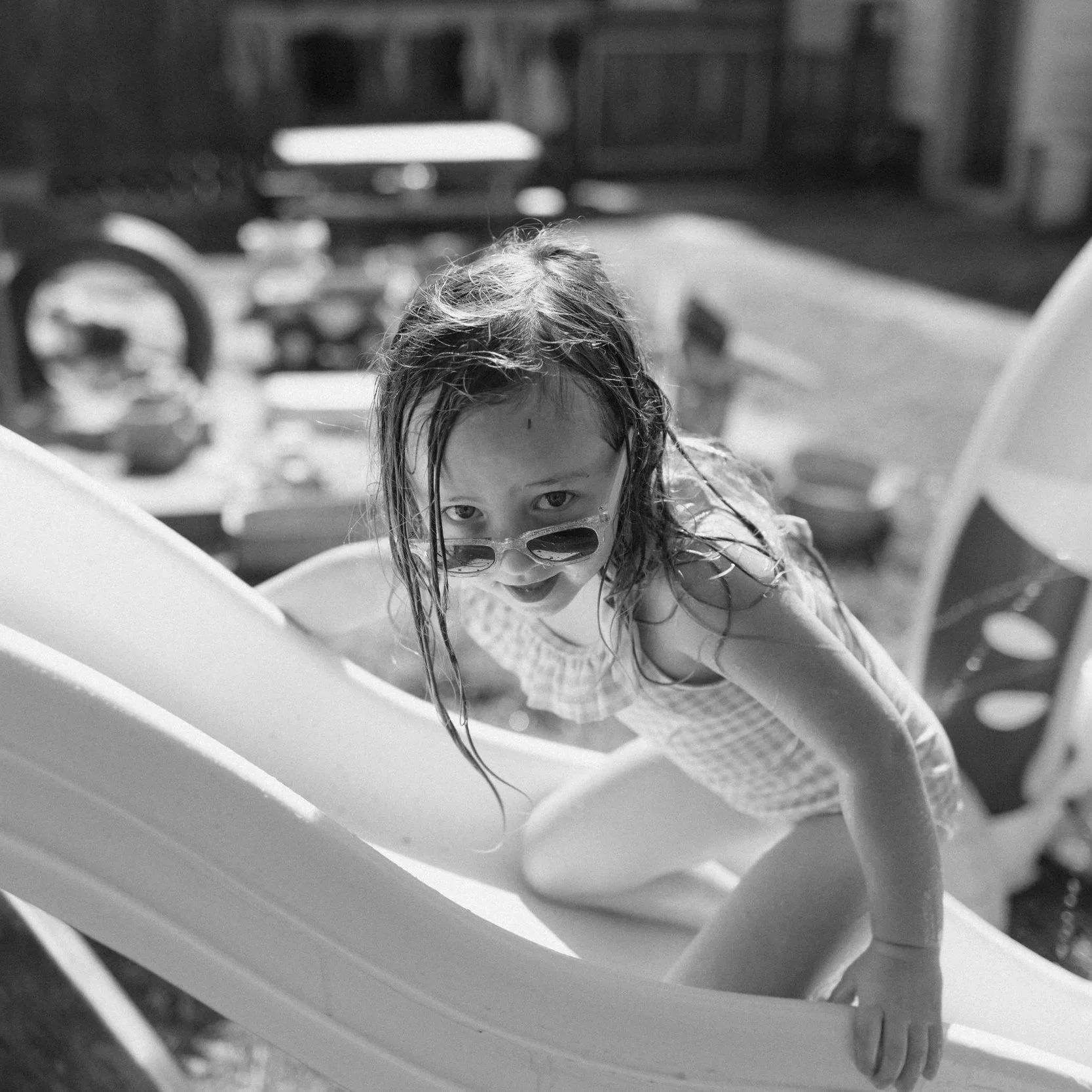 A young girl with wet hair and sunglasses, crawling on a playground slide, looking up at the camera in black and white.