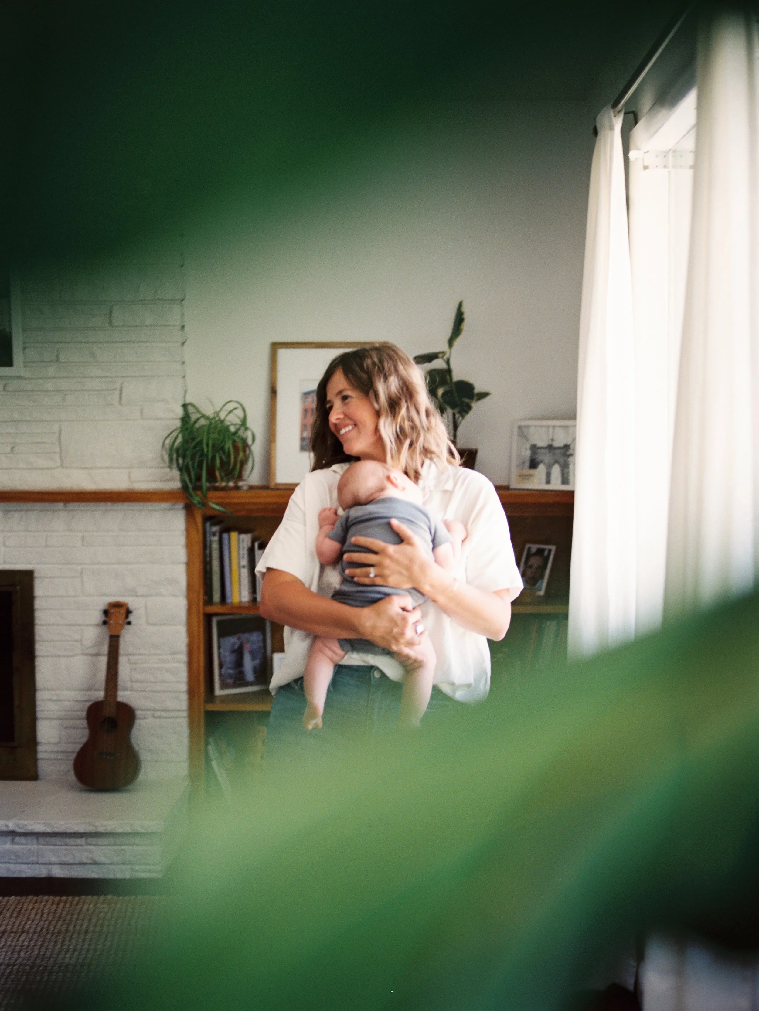 A woman with wavy brown hair smiling while holding a baby. They are inside a living room with a fireplace, a guitar, plants, and framed pictures. The photo is taken through green leaves or plants in the foreground.
