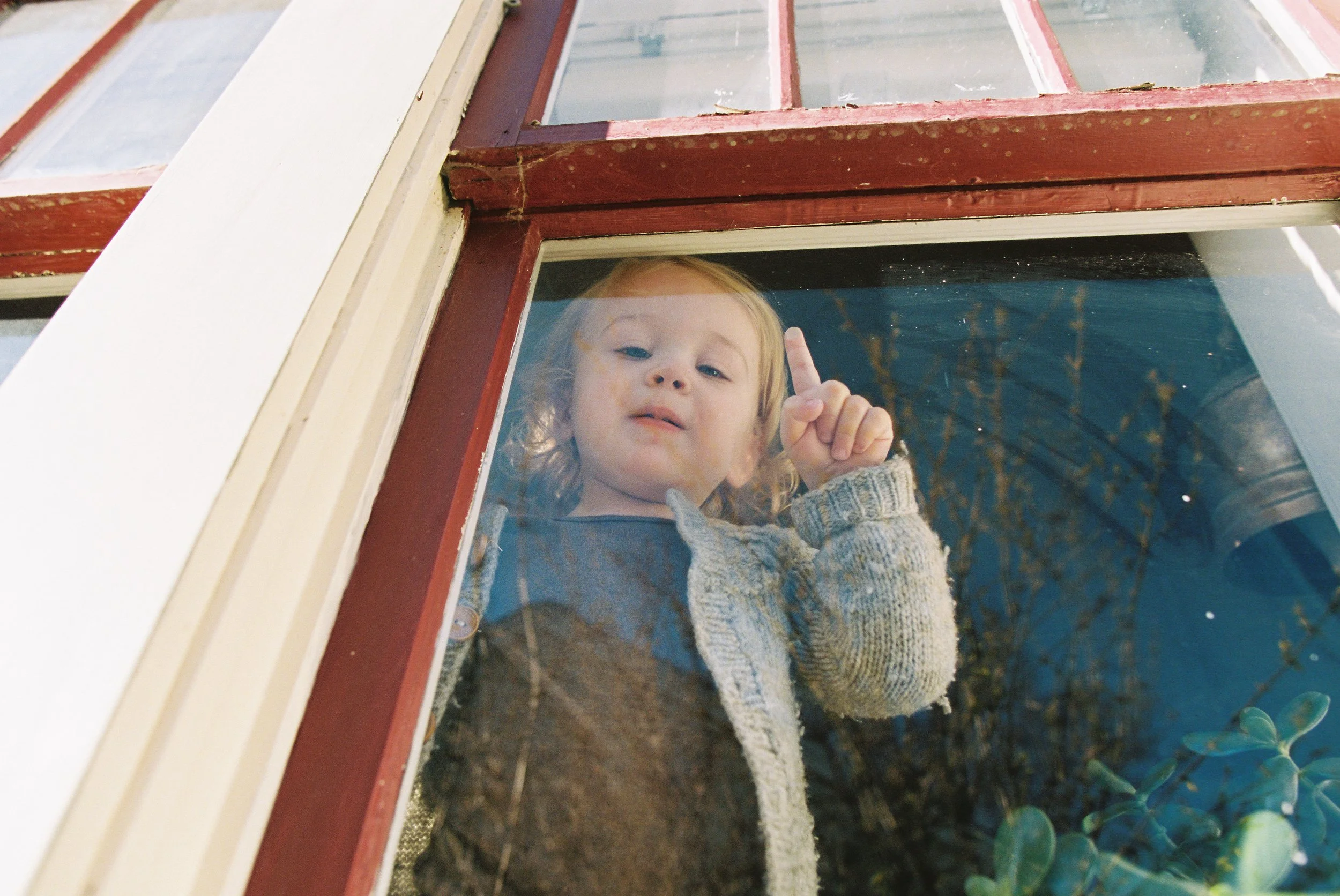 A young child with curly hair, wearing a gray knitted sweater, looking down through a window with red and beige framing, and pointing upward with their finger.