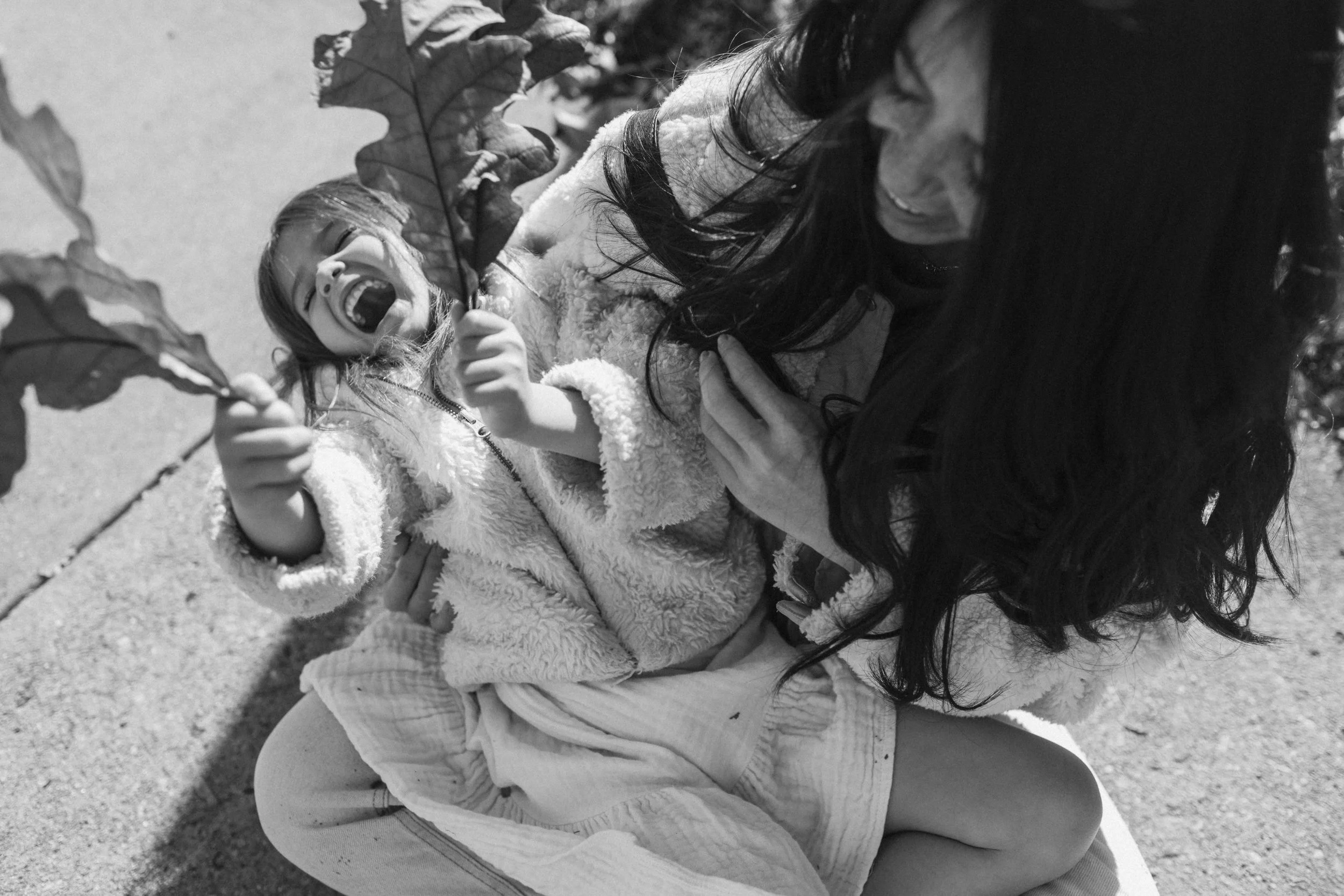 A woman and a young girl laughing and playing with large leaves outdoors.
