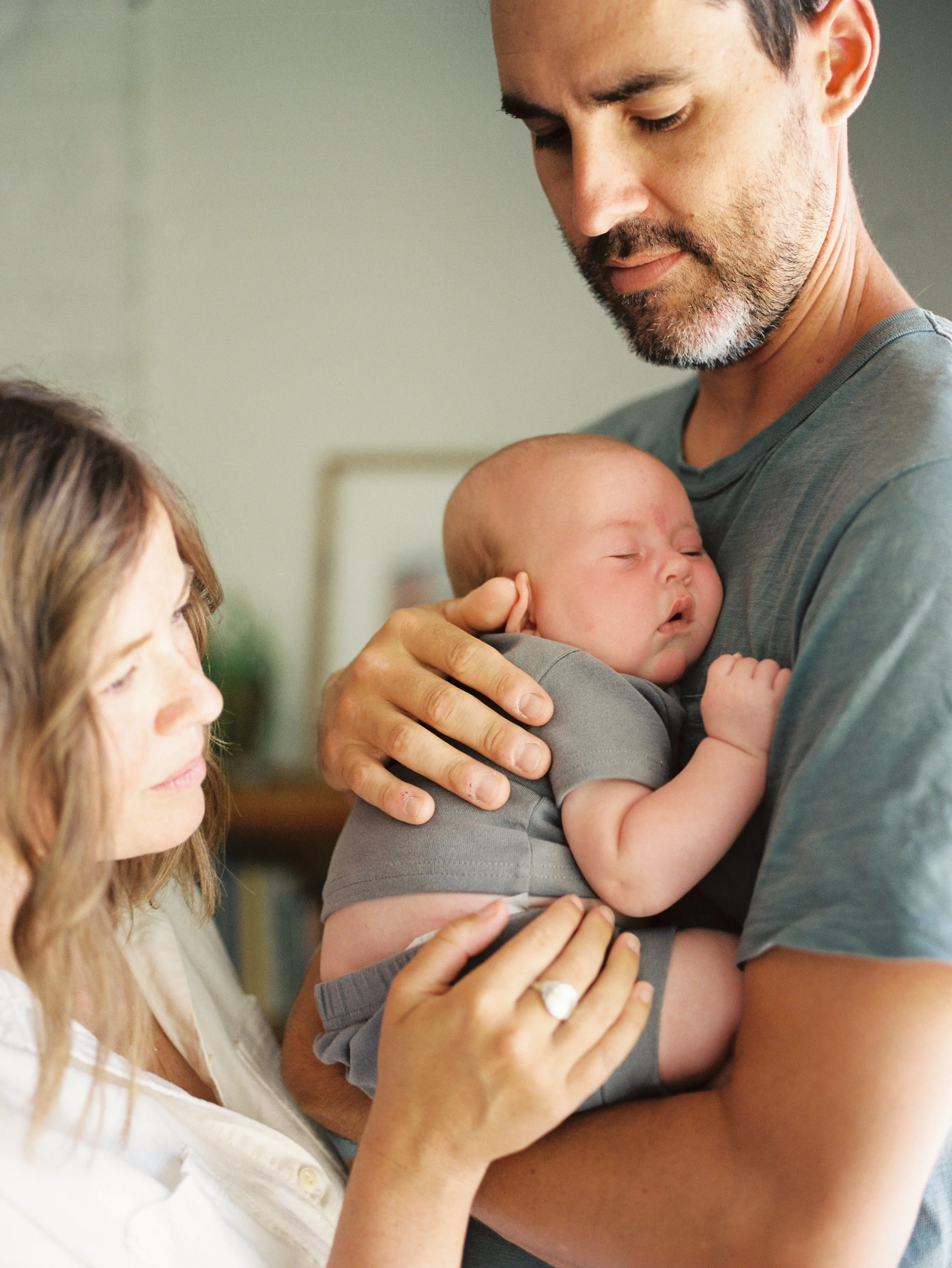 A man holding a sleeping baby while a woman gently touches the baby, in a cozy indoor setting.
