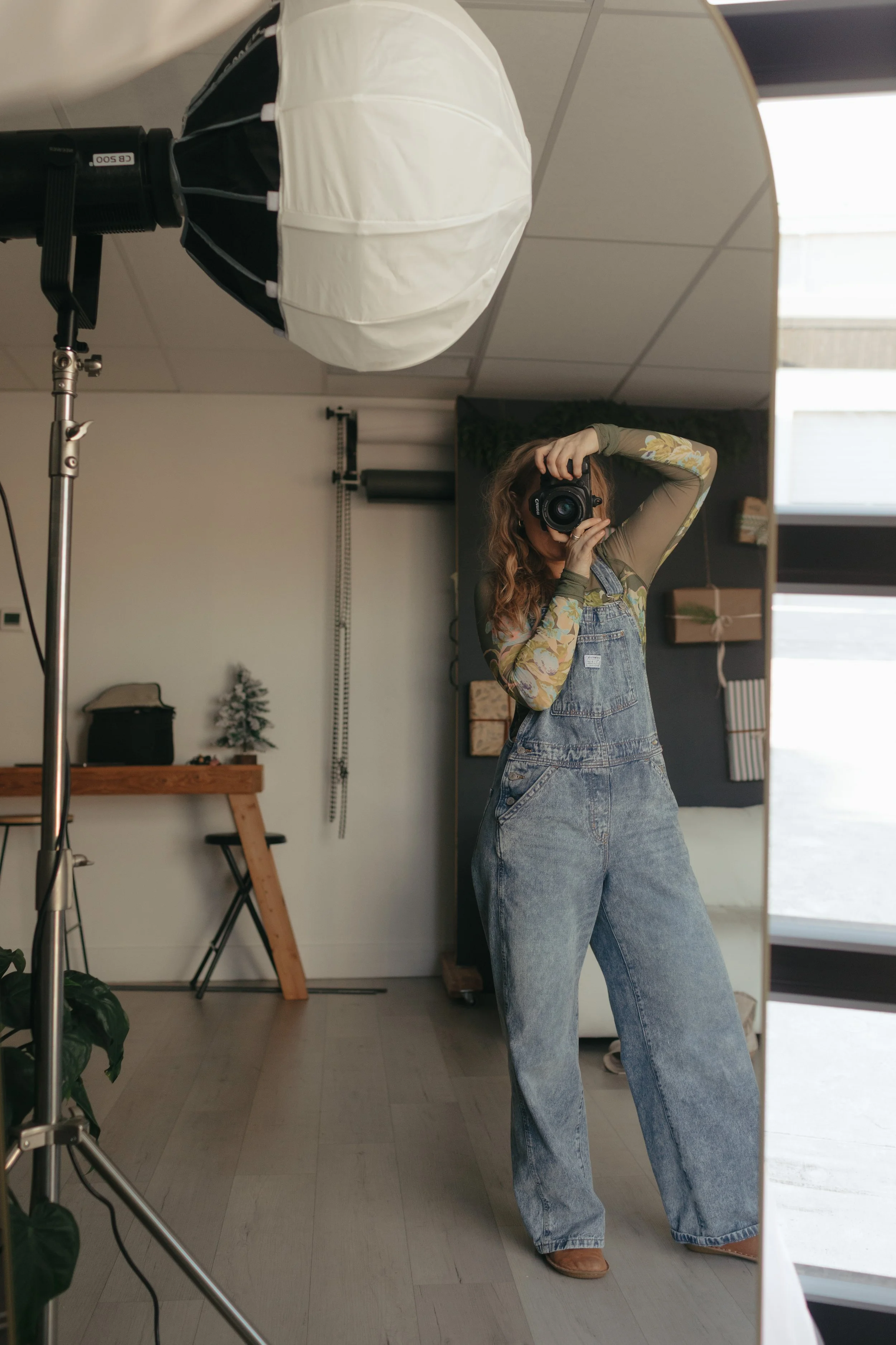 A woman in wide-legged jeans and a floral long-sleeve top taking a selfie in a mirror with a camera, in a room with a photography lighting setup.
