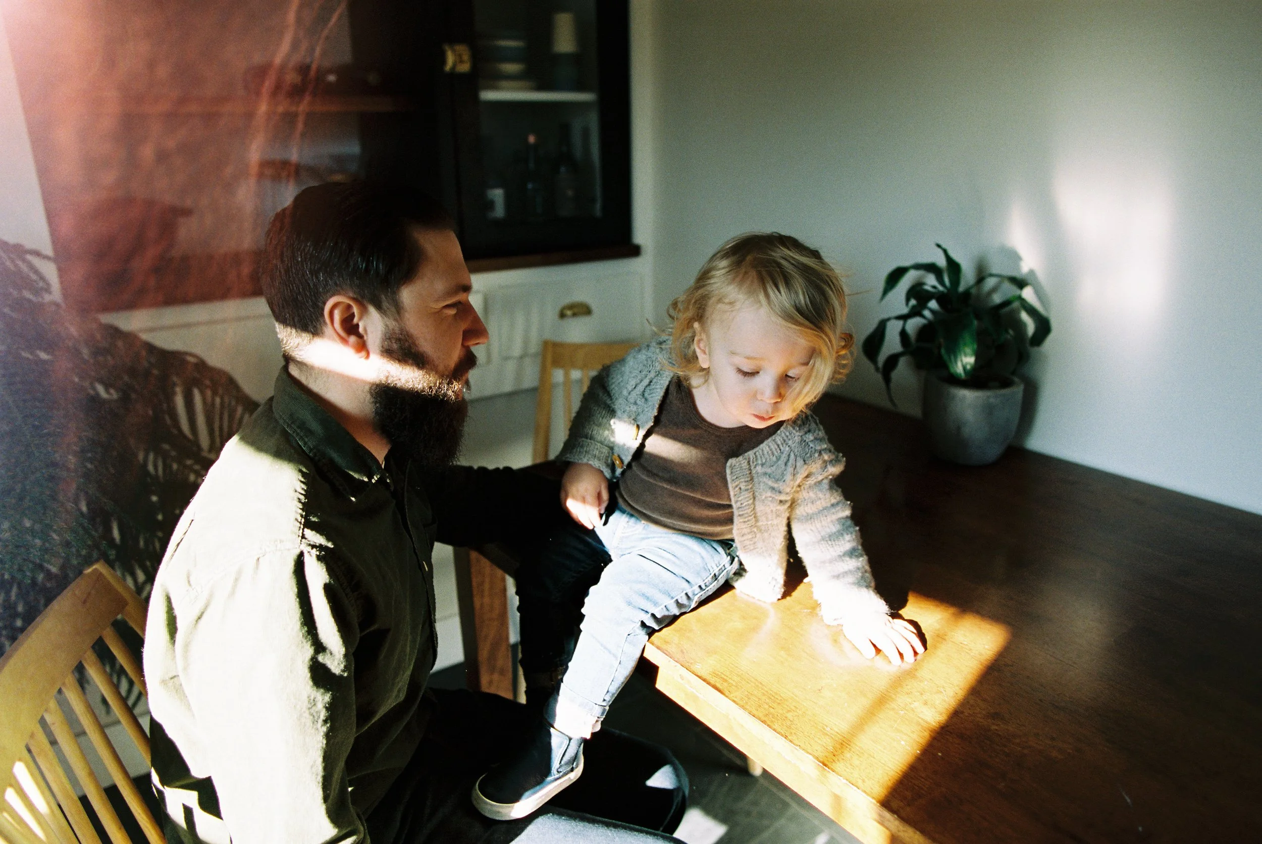 A man helping a young girl climb onto a wooden table in a sunlit room with a plant in a pot in the background.