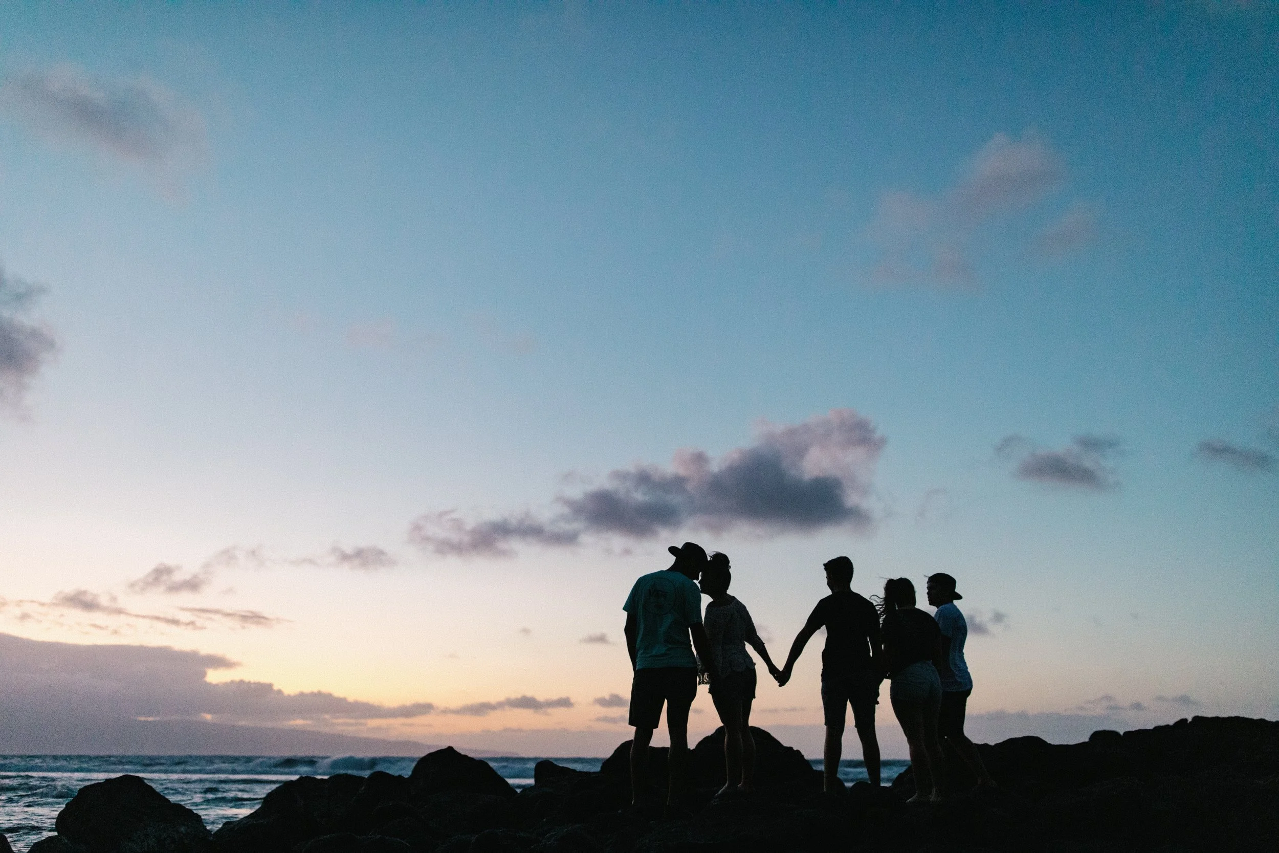 Silhouettes of six people holding hands on rocks by the ocean at sunset with clouds in the sky in Maui Hawaii