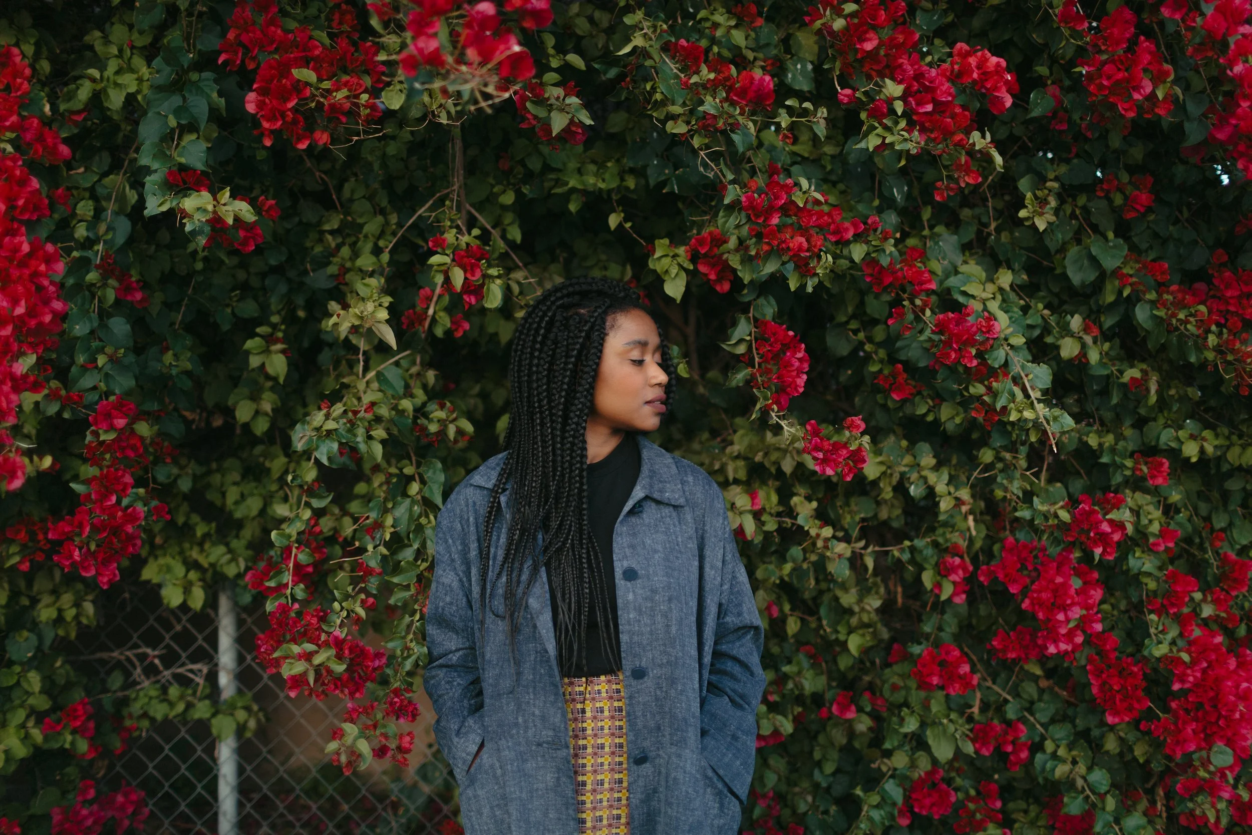 A woman with long, braided hair wearing a gray coat and plaid pants stands in front of a lush bush with bright red flowers and green leaves.