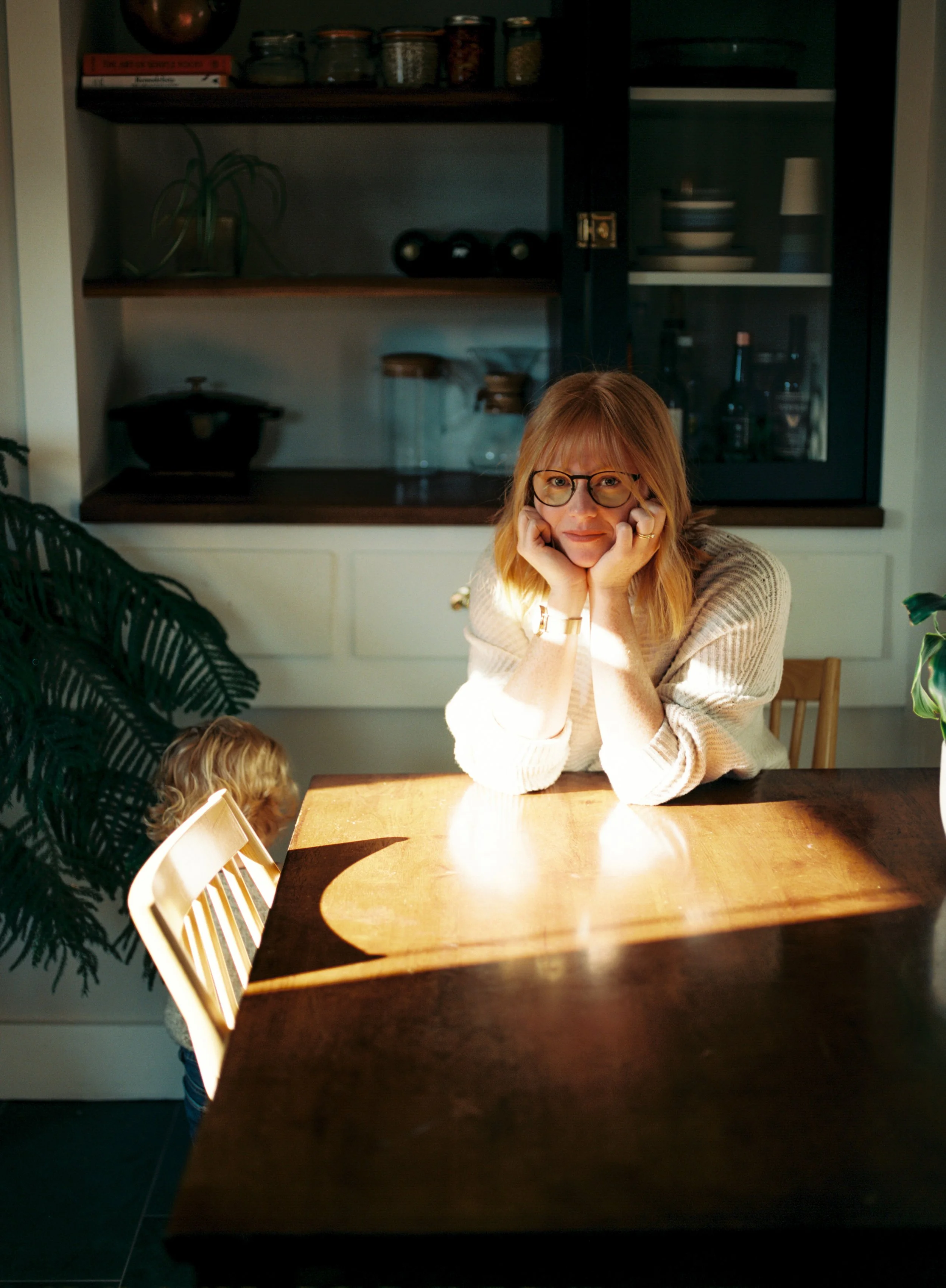 A woman with glasses and blond hair rests her chin on her hands, sitting at a wooden table with sunlight casting a shadow across it. A child with curly hair sits nearby, partially visible. The background features a kitchen with dark shelves, glass ja