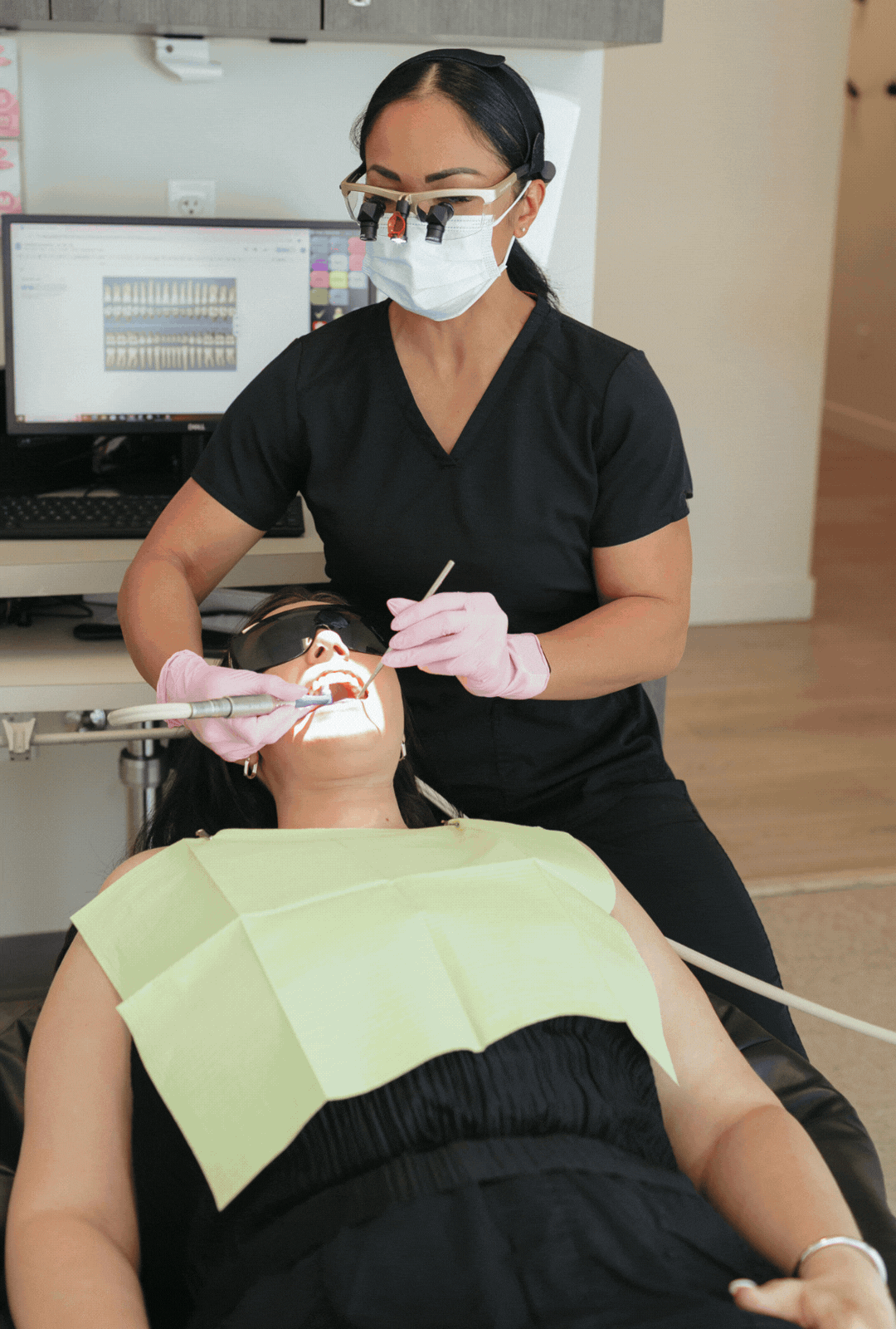 A dentist in black scrubs and a face mask performing a dental procedure on a patient lying back in a dental chair. The patient wears sunglasses and a dental bib, with dental tools in her mouth, and hearing protection on her ears.
