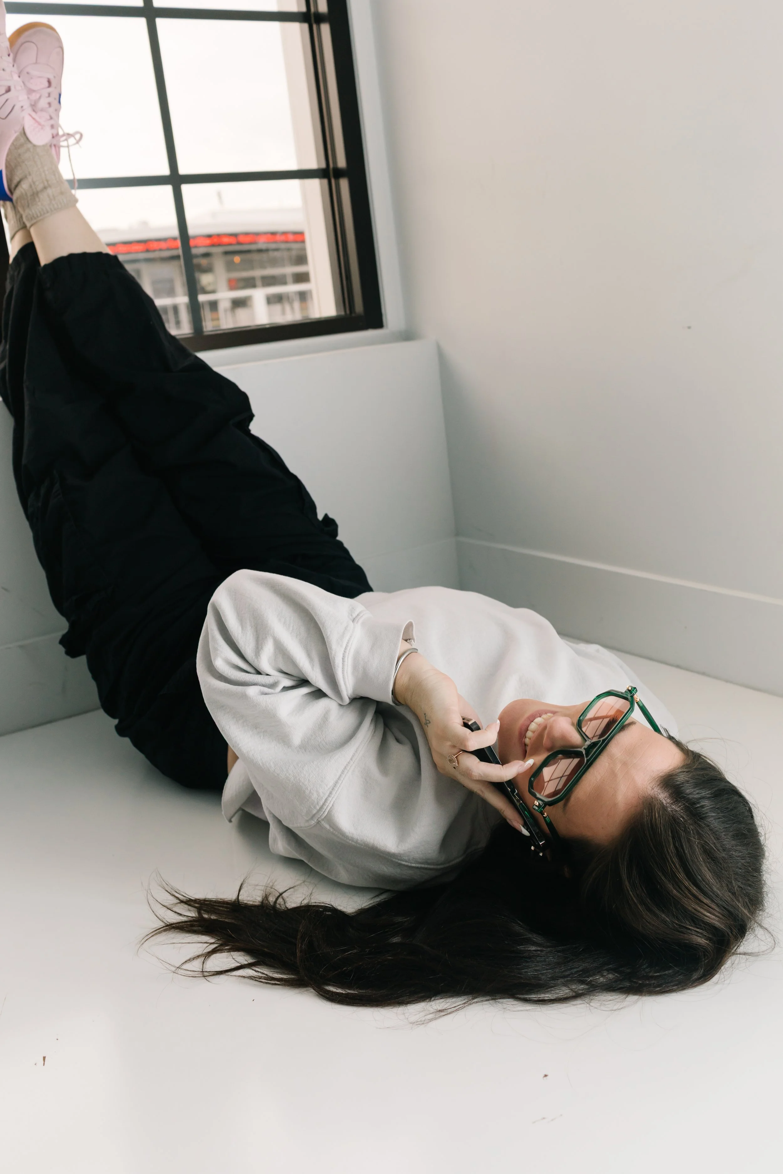 A woman with long dark hair and glasses is lying on the floor, smiling and talking on her phone, near a window with a city view.
