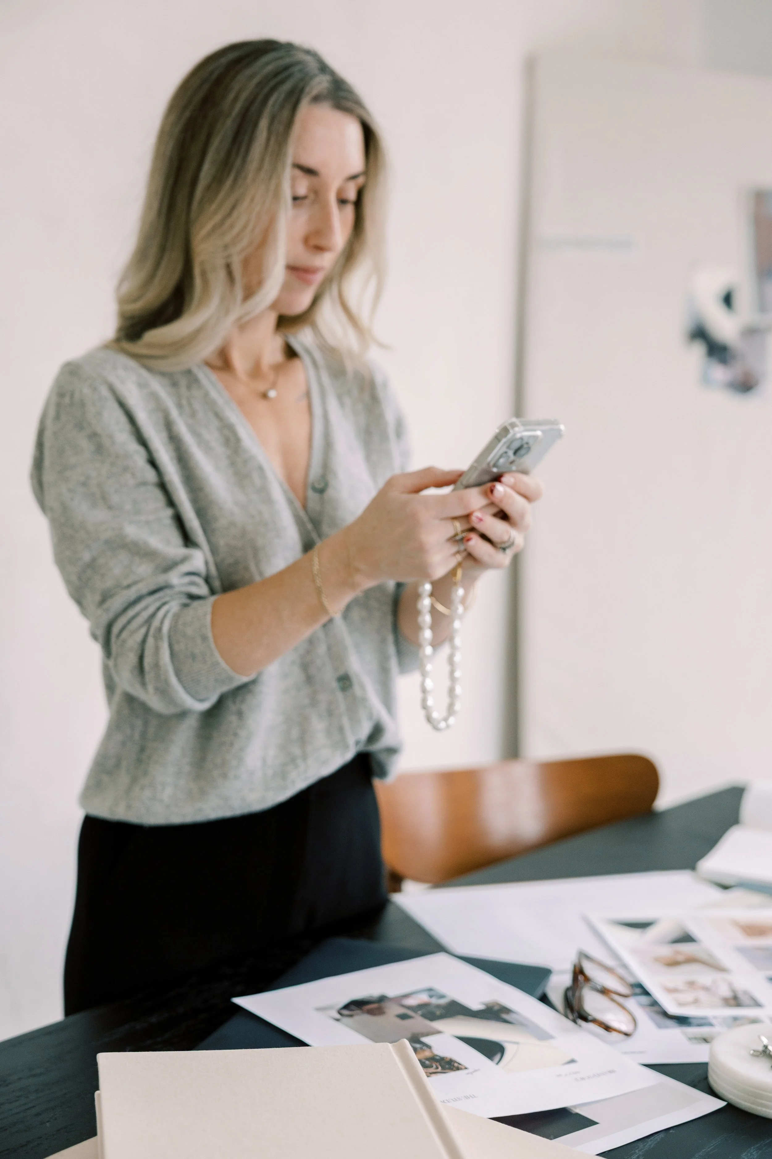 A woman in a gray sweater and black pants standing at a table, looking at her phone with a pearl necklace dangling from her hand, with papers, photos, and glasses on the table.