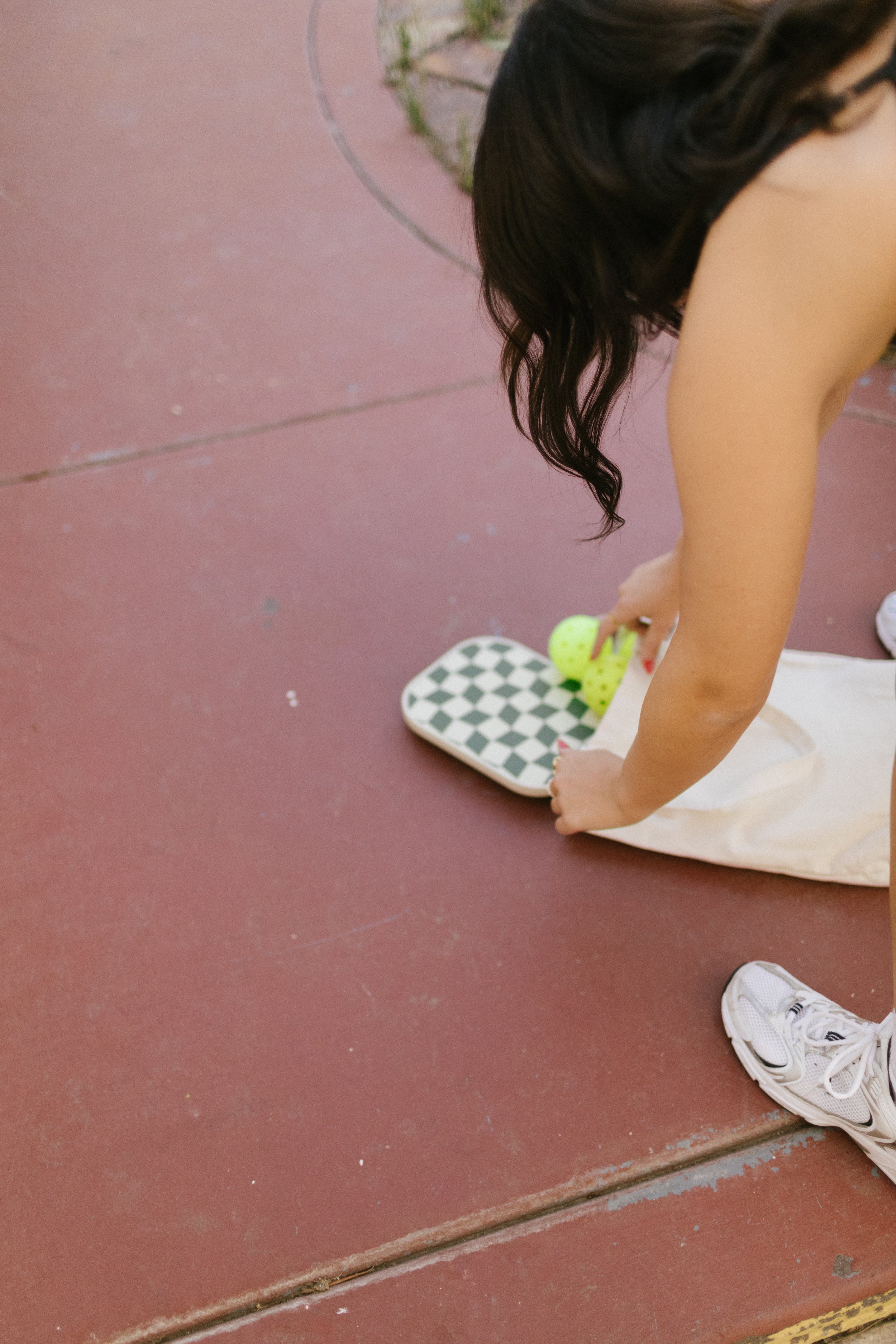 A woman is bending down on a red sidewalk next to a small paper bag, holding a yellow tennis ball, with another tennis ball inside the bag. She is wearing white sneakers and has long dark hair.