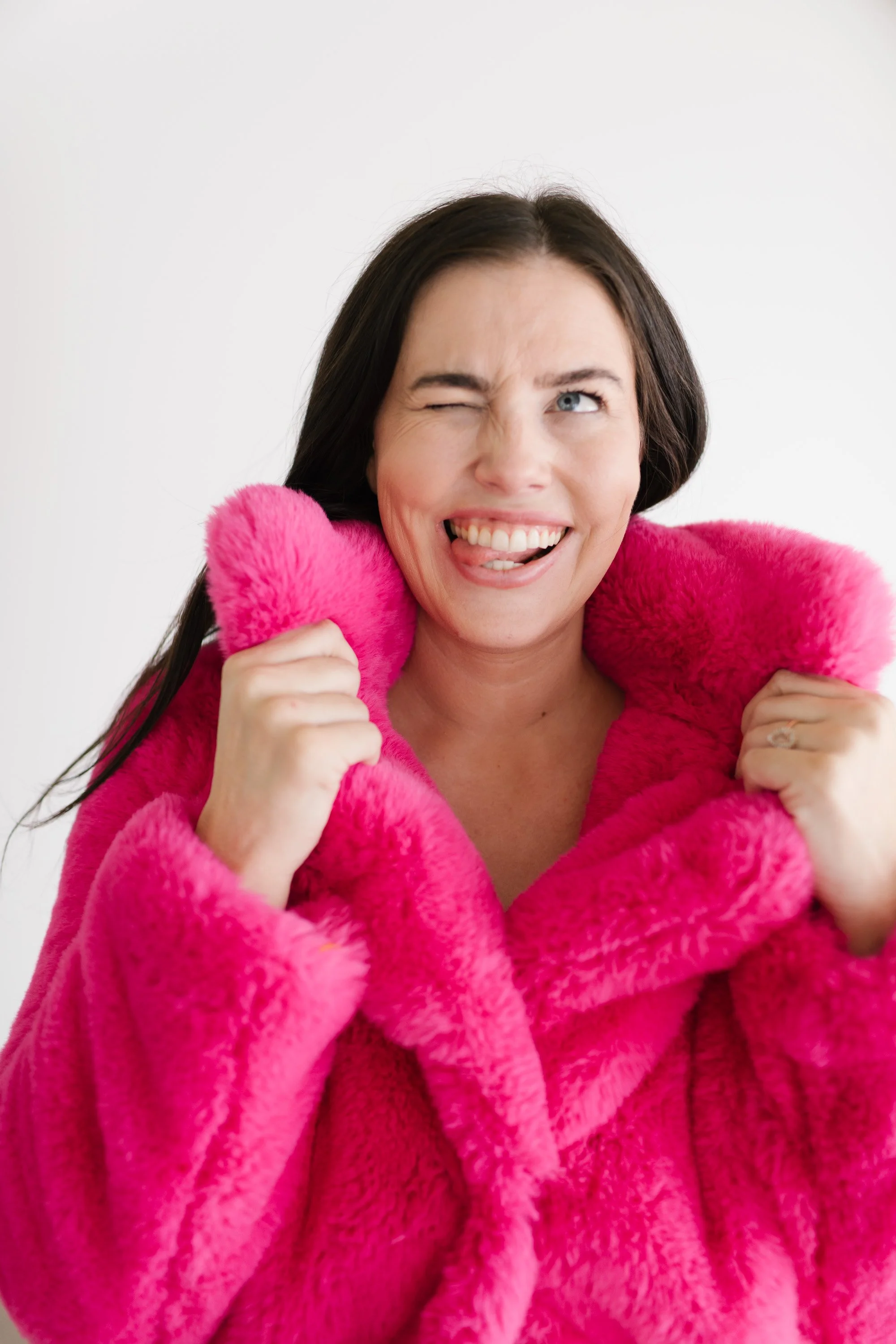 A woman with dark hair making a playful expression, winking and sticking out her tongue, holding a pink fuzzy coat around her shoulders against a plain white background.