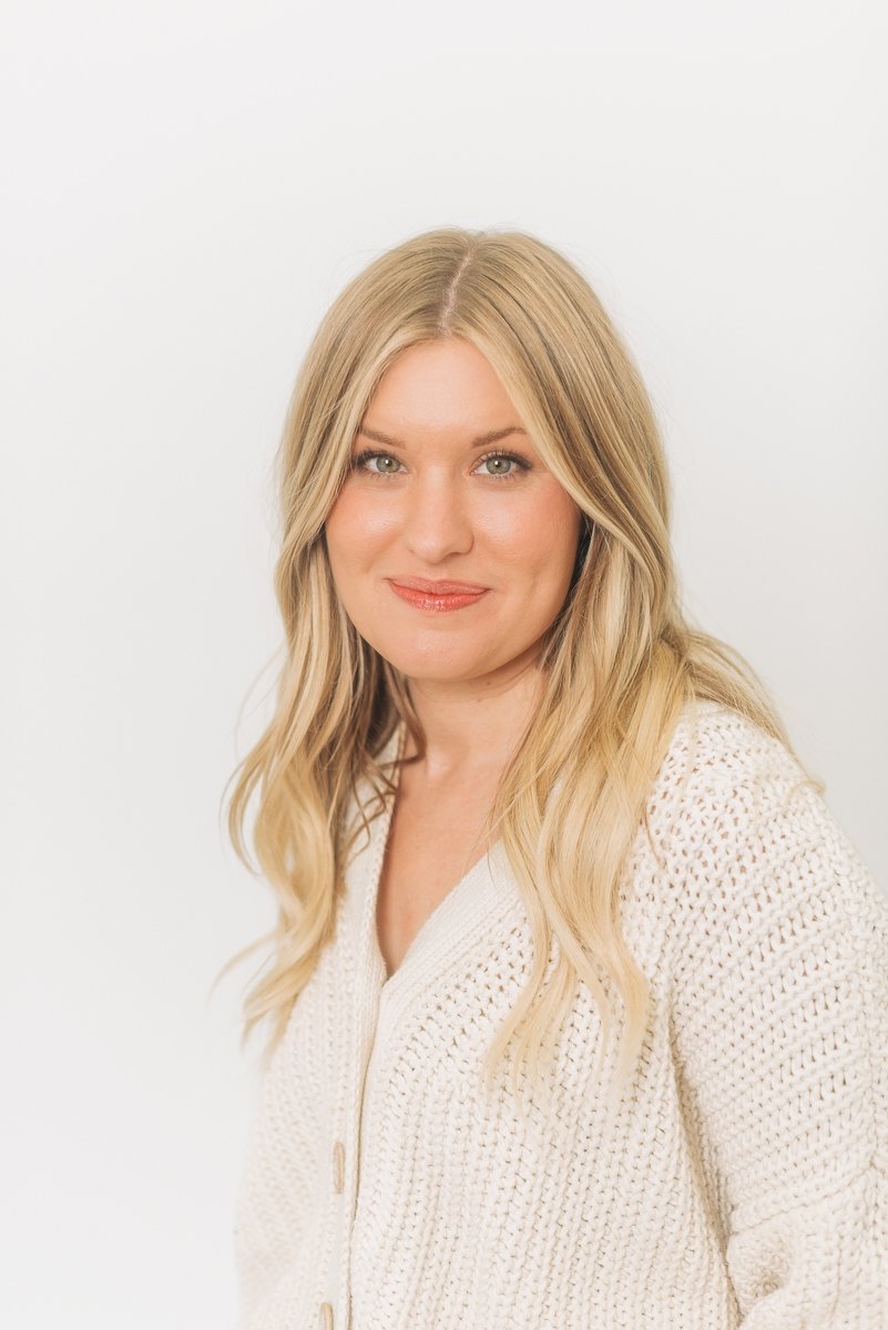 A woman with long blonde hair wearing a cream-colored knitted sweater smiling against a plain white background.
