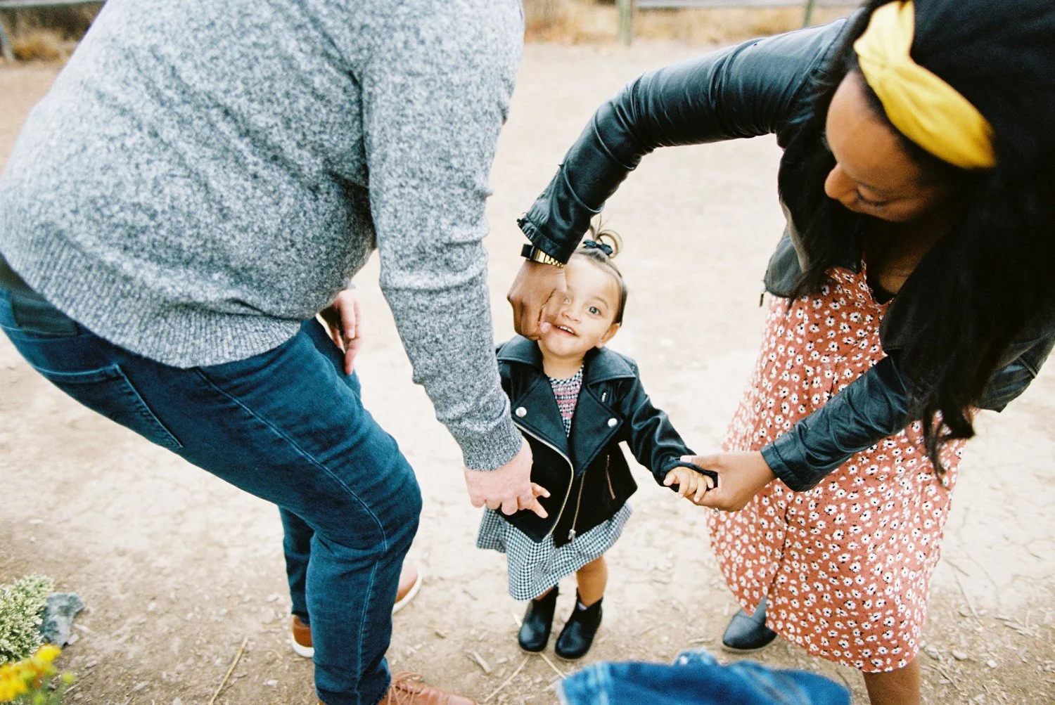 A young girl in a black leather jacket and black boots is holding hands with two adults, one on each side, in an outdoor setting in in Boise, Idaho.. The girl is looking up and smiling.