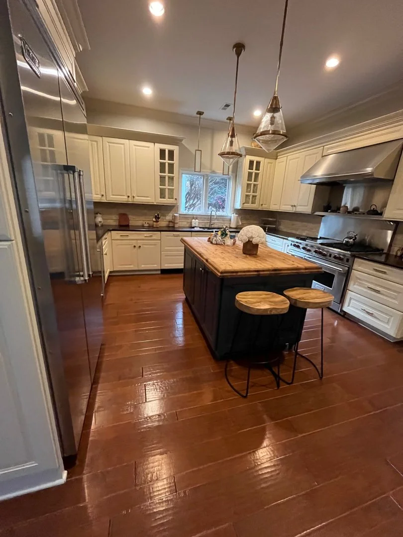 A spacious kitchen with white cabinets, a wooden island, and a hardwood floor. There are pendant lights hanging above the island, a window with a view of trees, and stainless steel appliances, including a refrigerator and a range hood.