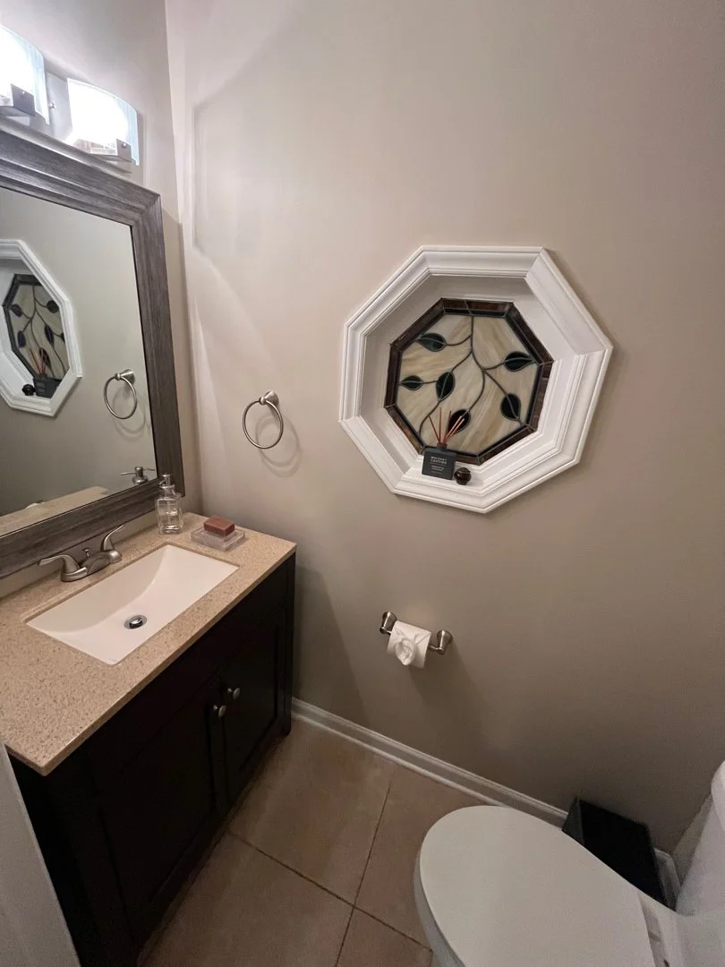 Small bathroom with a beige countertop vanity, dark wood cabinet, mirror, wall-mounted towel ring, octagonal stained glass window with a white frame, toilet paper holder with roll, and a white toilet.
