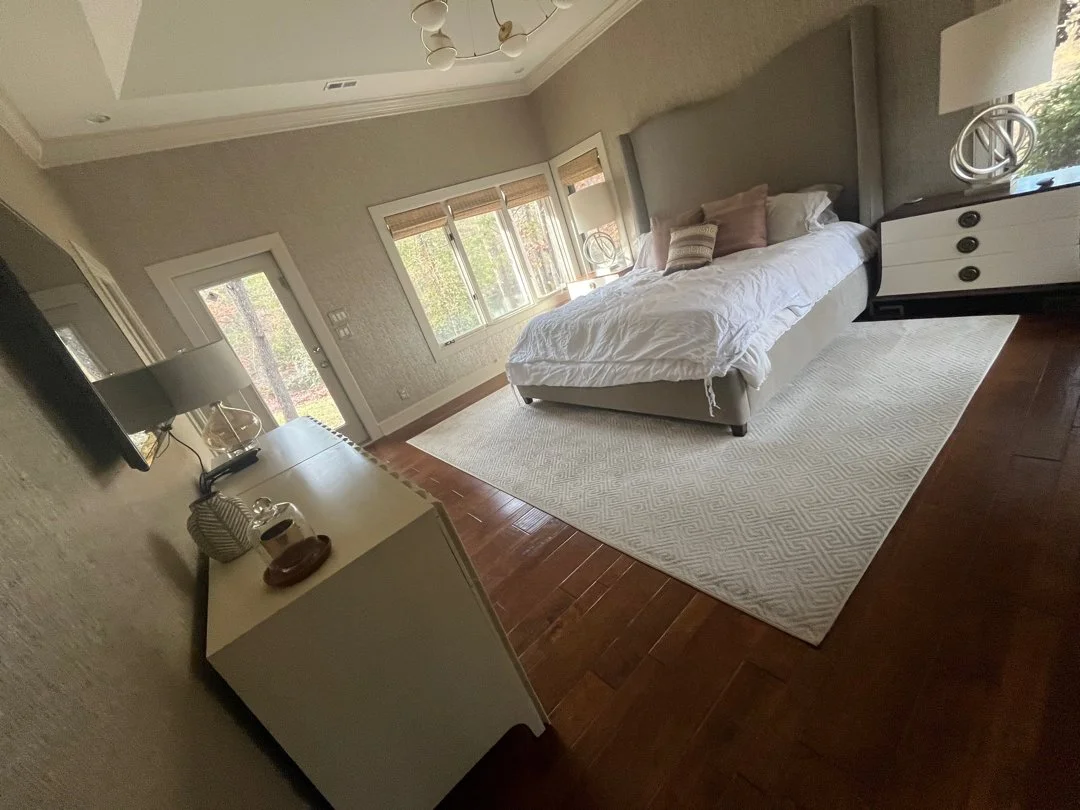 A bedroom with a bed, nightstand, and dresser, with windows showing a view of trees outside.