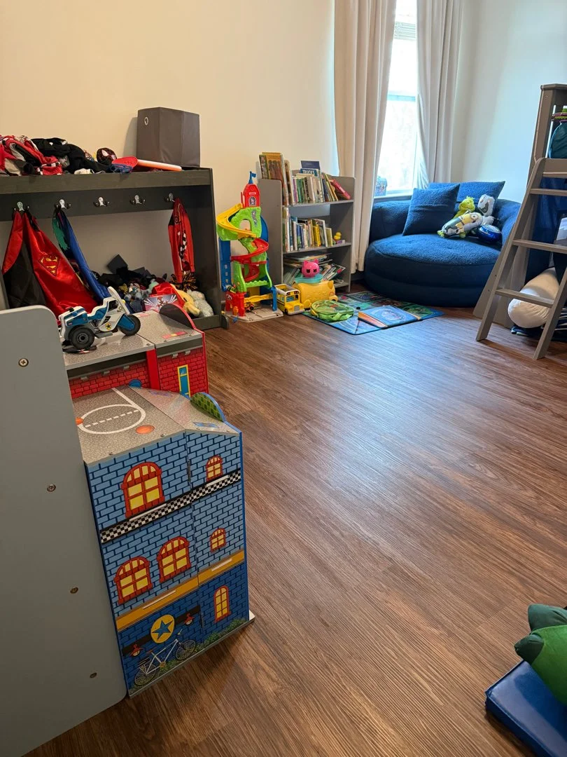 A child’s playroom with a blue couch, toys, books, a mini basketball court, and a large window with curtains.