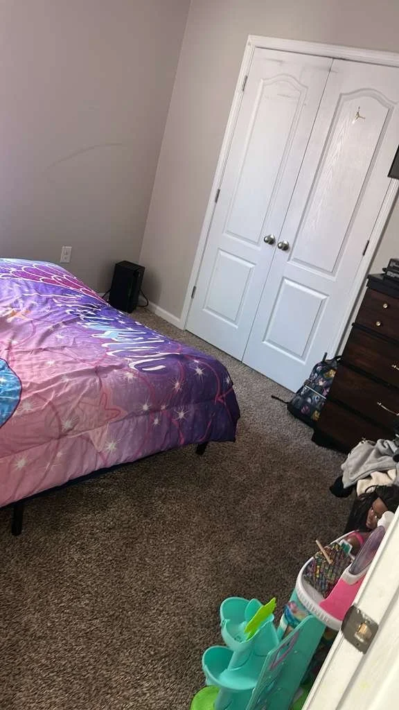 A bedroom with a bed covered in a purple and pink spaceship-themed comforter. There is a white closet door, a dark wooden dresser, and a small black speaker on the carpeted floor. A plastic toy organizer with compartments is visible near the doorway.