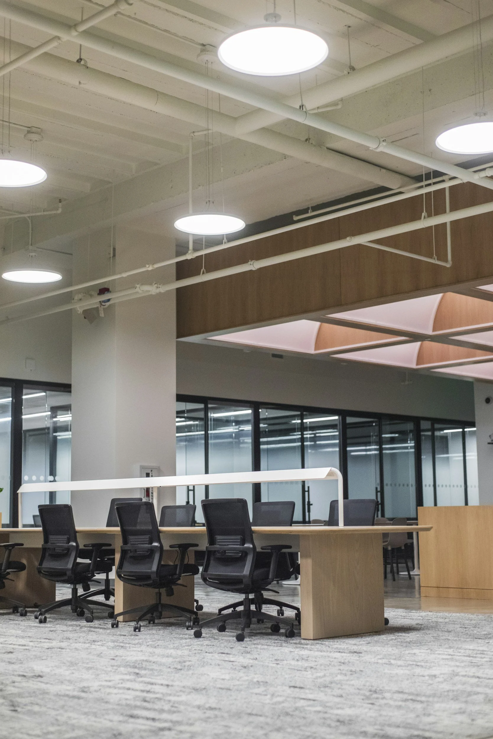 Modern conference room with a large wooden table, black office chairs, bright overhead lighting, and glass wall partitions.