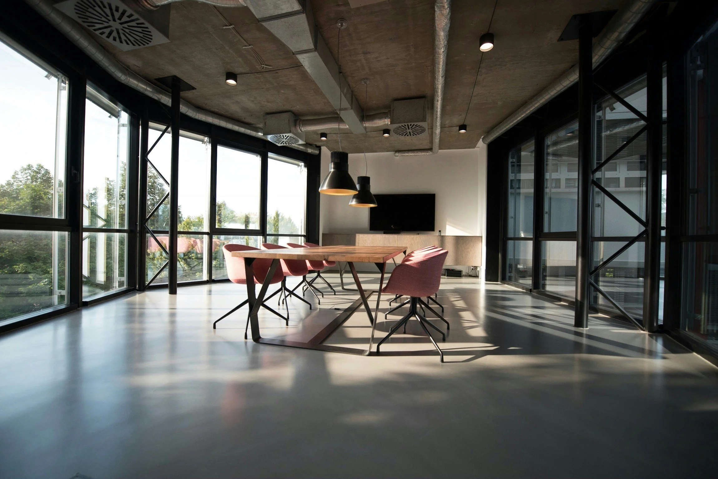 Modern office conference room with a wooden table surrounded by pink chairs, large windows with a city view, and a wall-mounted television, illuminated by hanging pendant lights.