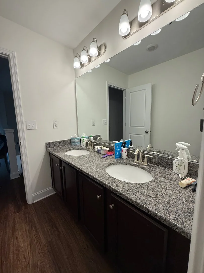 A bathroom vanity with a granite countertop, two sinks, dark wooden cabinets, and a large mirror with four overhead lights. The countertop has various toiletries and cleaning supplies on it.